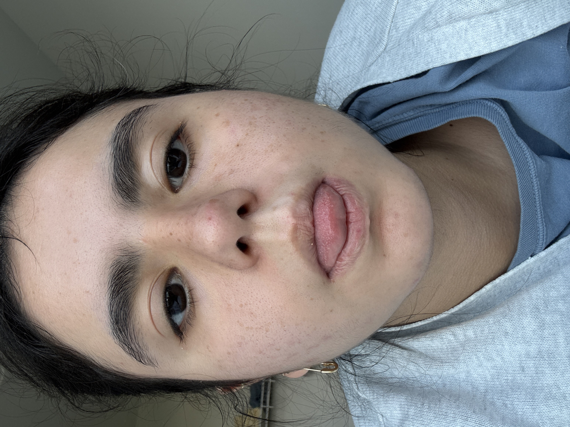 A close-up, rotated portrait of a person, likely female, is captured indoors in Austin, United States of America. The subject's head is tilted, and they are looking directly at the camera with a playful expression, their tongue partially extended and pressed against their upper lip.
The person has dark, slightly disheveled hair, dark, prominent eyebrows, and dark brown eyes. Their skin displays visible pores and a scattering of freckles or small moles across the forehead and cheeks. Their lips appear somewhat dry or chapped. They are wearing a light blue crew-neck t-shirt underneath a light grey hooded garment, possibly a sweatshirt or jacket. A small, golden earring, shaped like a safety pin, is visible near their right ear.
The background is softly blurred and appears to be a plain, light grey-green color, suggesting an indoor setting. The lighting is soft and even, highlighting the facial features without harsh shadows. No text is visible in the image.