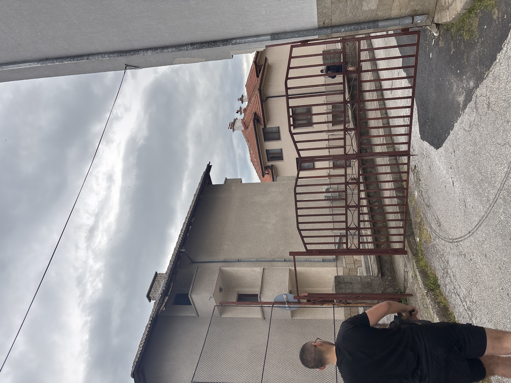 A low-angle shot of an alleyway in Zlatograd, Bulgaria. Buildings stand to either side, partly obscuring the cloudy sky. An open gate and a black fence are visible. A man in a black shirt and shorts stands in the foreground. The pavement is grey.
