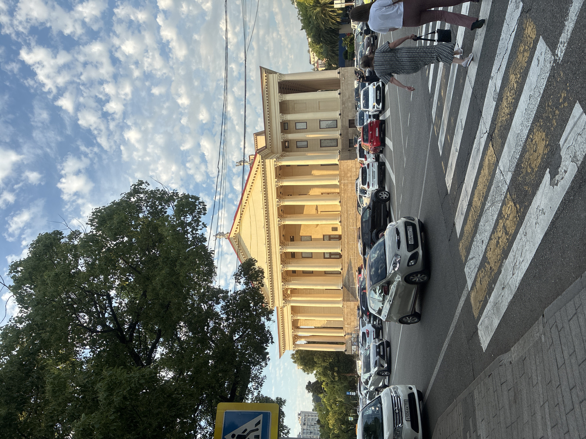 The image depicts a street scene in Sochi, Russia, with a large, neoclassical building featuring prominent columns. A busy street runs in front of the building, lined with parked cars. Pedestrians are crossing a zebra-striped crosswalk in the foreground. One pedestrian, a woman in a patterned dress, is mid-crosswalk, holding a handbag. Another person, wearing a white t-shirt and maroon pants, walks behind her.
The time of day appears to be late afternoon or early evening, with the sun casting long shadows and illuminating the building with a warm glow. The sky is partly cloudy, with scattered white clouds against a blue backdrop. Lush green trees, including a large, full tree on the left side of the frame, suggest a pleasant climate.
Visible text includes license plates on some of the cars. For instance, one car has the plate "334 OXY 90". Another car's plate reads "E506 AX 193". A blue and white traffic sign, likely indicating a pedestrian crossing or school zone, is also visible on the left. The overall atmosphere is one of urban activity on a clear day.