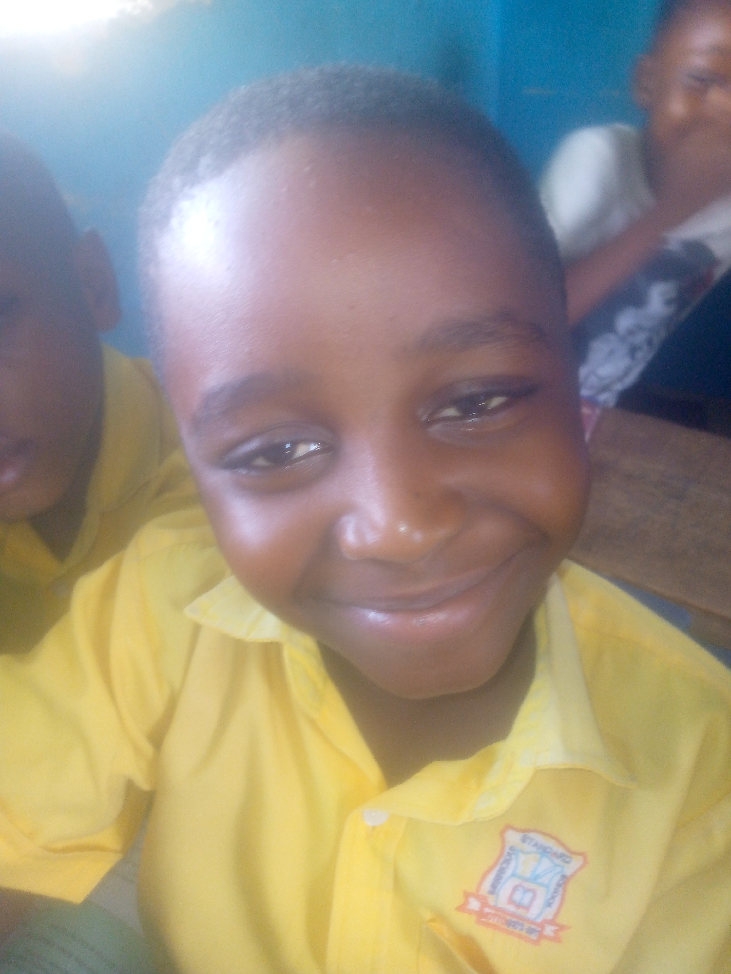 A close-up shot shows a young Black boy in a yellow collared shirt with short, dark hair. He is smiling slightly at the camera. Another child is partially visible behind him to the left, also wearing a yellow shirt. A third child can be seen in the background on the right, holding their hand to their mouth. The boy's shirt has a school emblem on it. The environment appears to be indoors, potentially a classroom setting. Though the city location is inferred from the prompt, the image itself does not reveal or depict the city of Ikorodu, Nigeria.