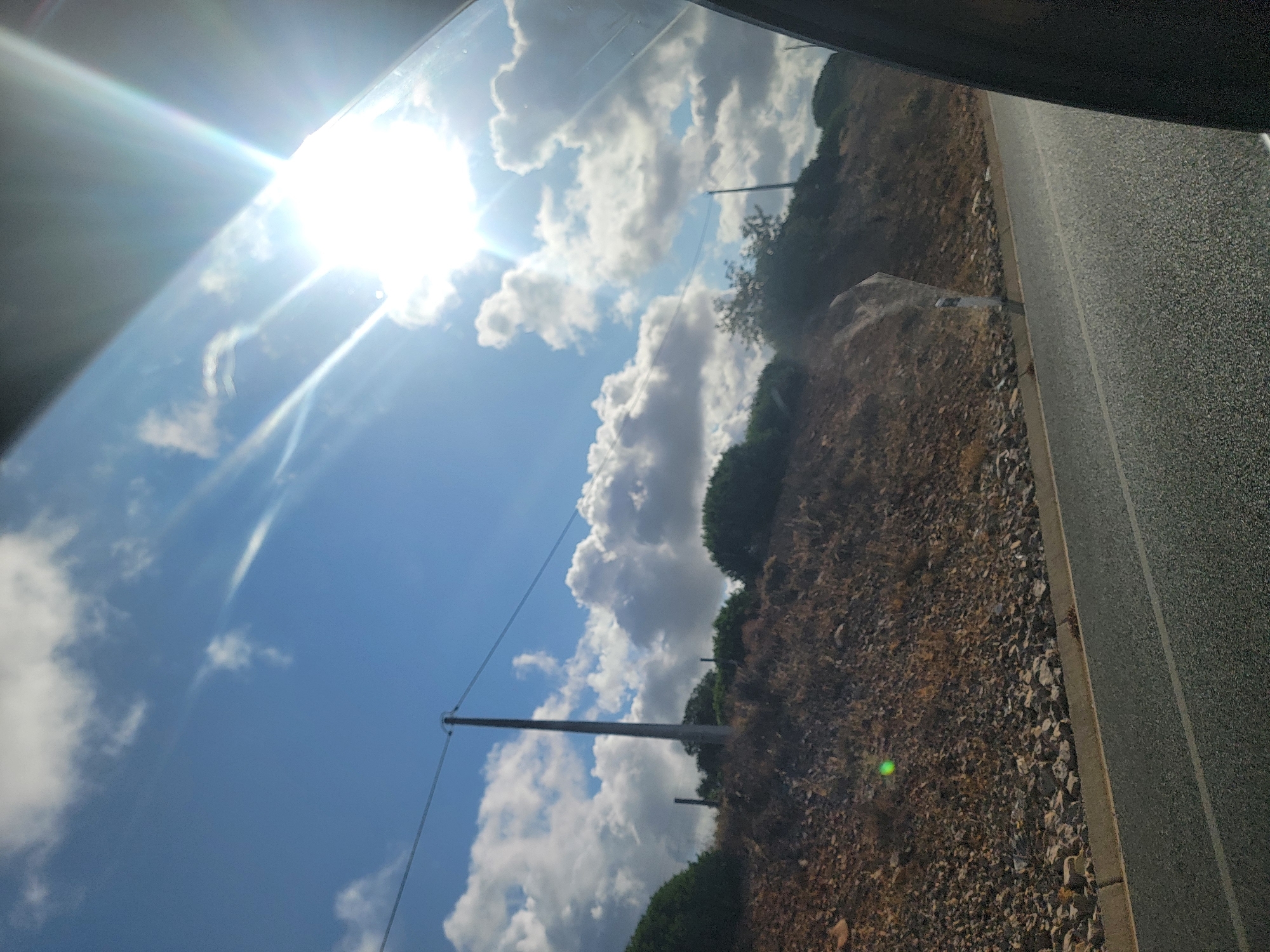 The image shows a roadside view from inside a vehicle, looking towards a sunlit sky over a scrub-covered embankment. The sky is a bright blue with fluffy white clouds. The sun is very bright, creating lens flare and rays of light, and it is partially obscured by the window frame on the right. There are telephone poles visible along the top of the embankment, with wires stretching between them. The embankment itself is covered in dry brush, rocks, and some scattered greenery. A paved road with a white line marking is visible in the foreground, with a concrete curb. A faint green circular light artifact is present on the embankment. The scene appears to be in Almancil, Portugal.