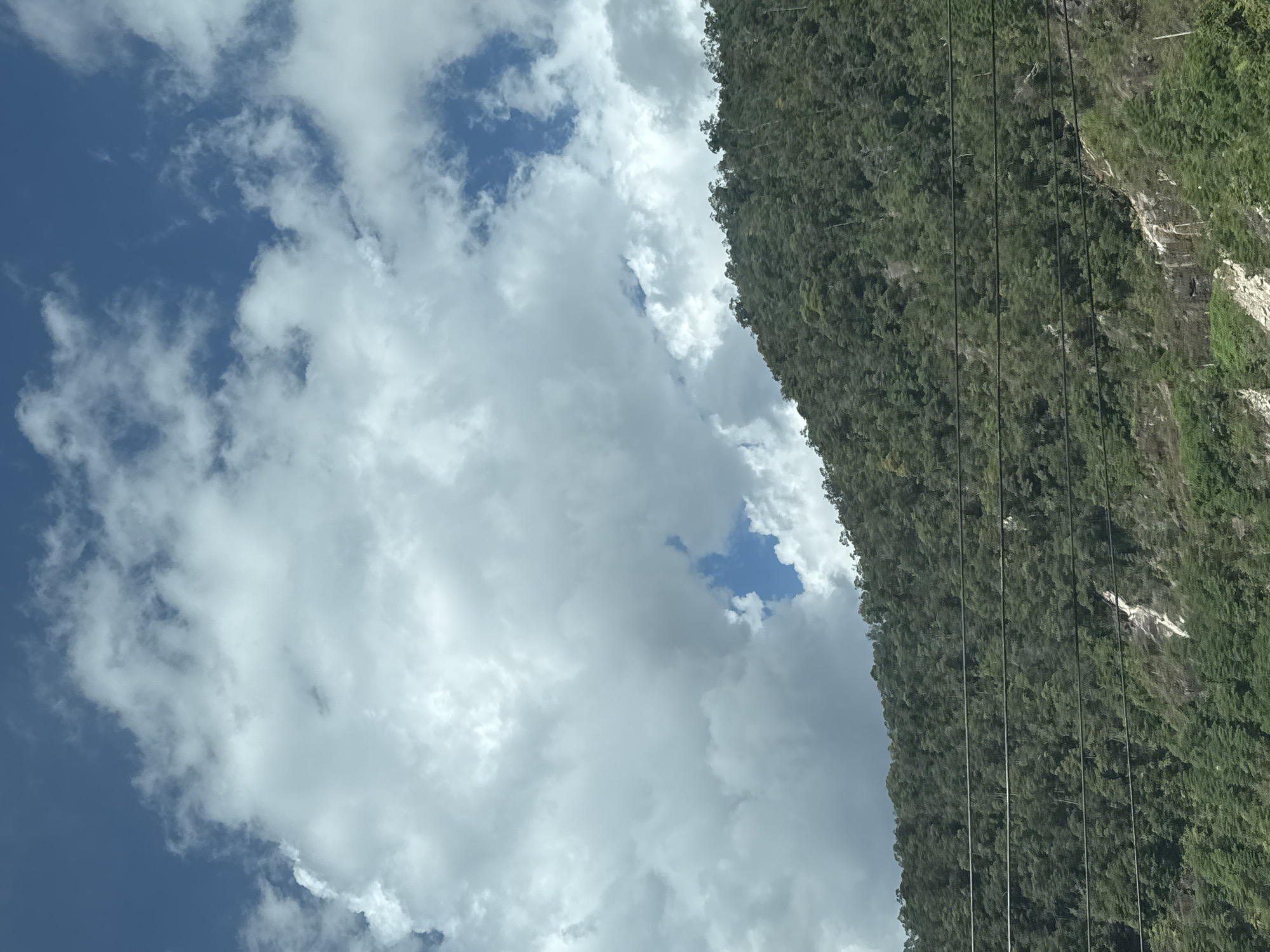 The image depicts a lush, green mountainside in Nam Khanh Vinh, Vietnam. Dense forest covers the slopes, interspersed with rocky outcrops and a small, white waterfall. Several dark power lines stretch horizontally across the mid-ground of the mountain. Above the mountain, a dynamic sky is visible, filled with a mix of bright blue patches and large, billowing white clouds. The scene is a natural landscape, devoid of any visible people or human activity beyond the power lines.