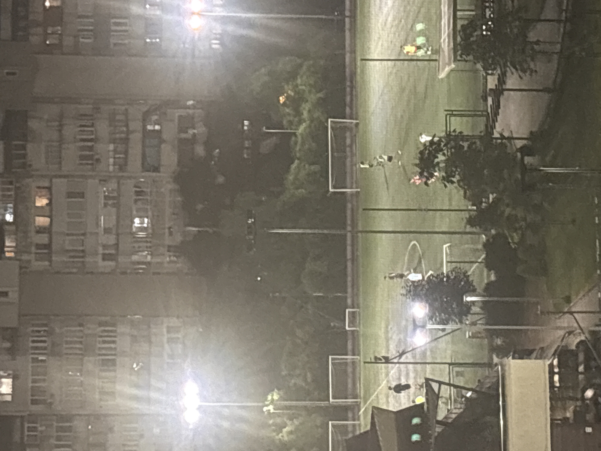An aerial, nighttime shot shows an outdoor soccer field with several players. The field is illuminated by overhead lights. In the background, a multi-story apartment building is visible. A few trees are scattered around the perimeter of the field. One person is standing on a raised platform to the left of the frame, which appears to be a small spectator area or press box.