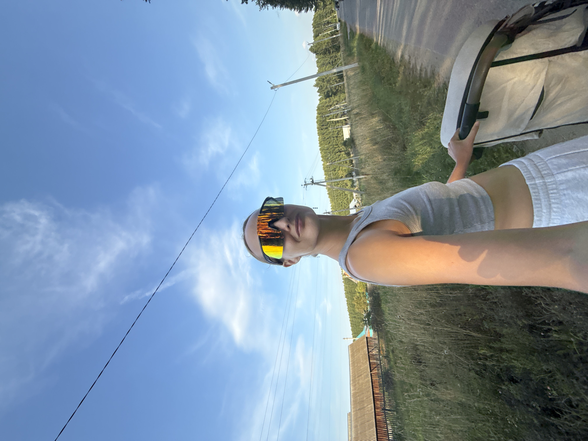 An individual is pictured from the chest up, holding the handle of a stroller with their right hand. They are wearing oversized, reflective sunglasses and a grey tank top. The background consists of a rural landscape with a dirt road, tall grass, wooden fences, and buildings, indicative of the village of Podshivalovo, Russia. Power lines stretch across the bright blue sky, which is dotted with wispy white clouds. The lighting suggests it is late afternoon.