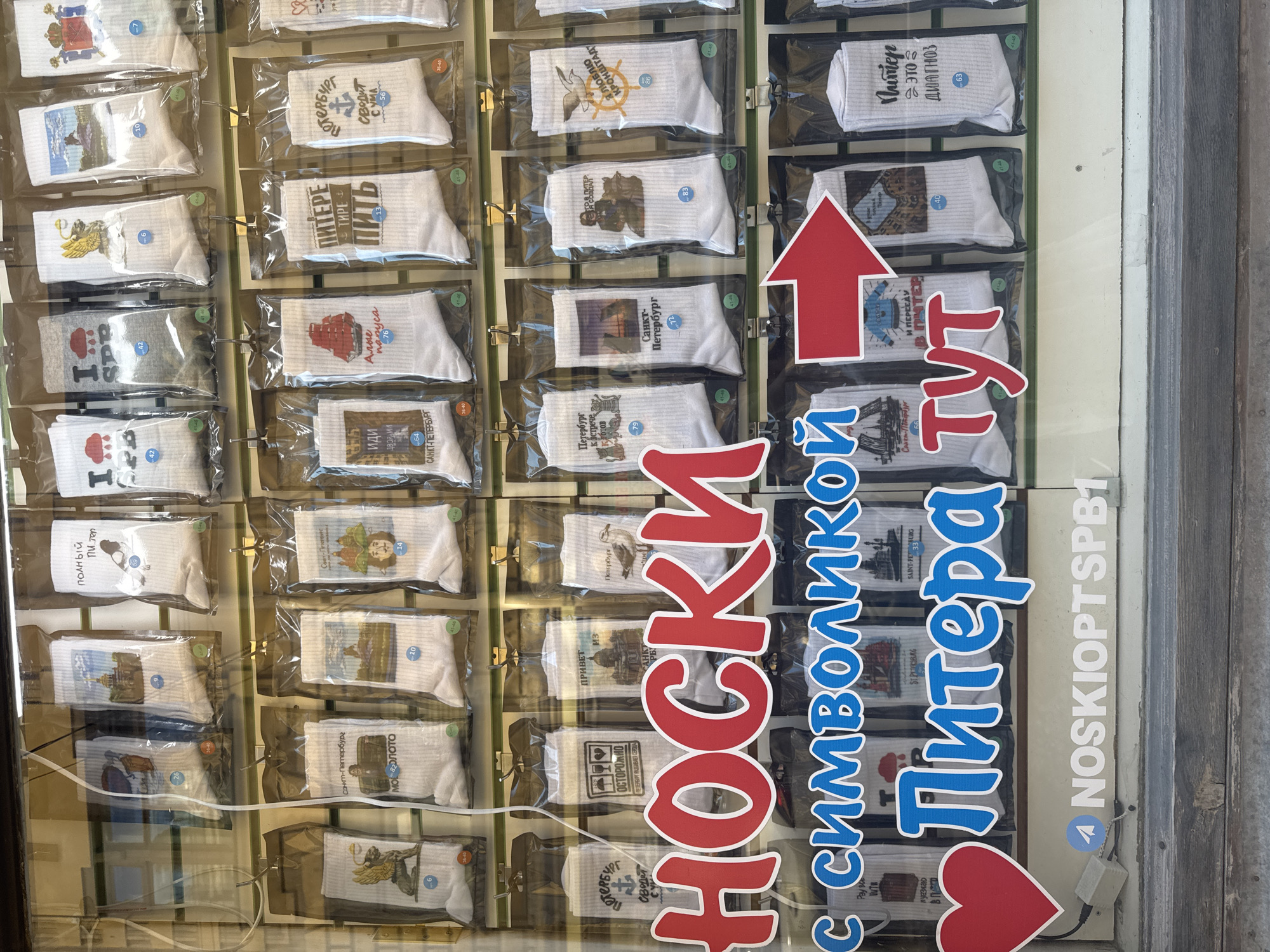 This is a close-up, eye-level shot of a shop window display featuring numerous pairs of white socks packaged in clear plastic. The socks are adorned with various images and text, many of which appear to be related to Saint Petersburg, Russia, indicated by the prominent Cyrillic text "НОСКИ С СИМВОЛИКОЙ Питера тут" (Socks with Petersburg symbols here).
The socks are arranged in rows on hangers behind a glass pane. The top row displays socks with images of landmarks, historical scenes, and humorous graphics. The second row continues this theme, with socks featuring notable Saint Petersburg motifs like the Bronze Horseman, St. Isaac's Cathedral, and the iconic "Peter the Great" imagery. The third and fourth rows also showcase socks with Saint Petersburg-themed designs. A large red heart graphic is positioned on the left side of the frame, below the "НОСКИ" text. Below the display of socks, a Telegram logo and the handle "NOSKIOPTSPB1" are visible, suggesting an online presence or ordering information. The background shows a reflection of buildings with windows, hinting at the external environment. The overall impression is a retail display designed to attract customers interested in souvenirs or themed apparel related to Saint Petersburg.