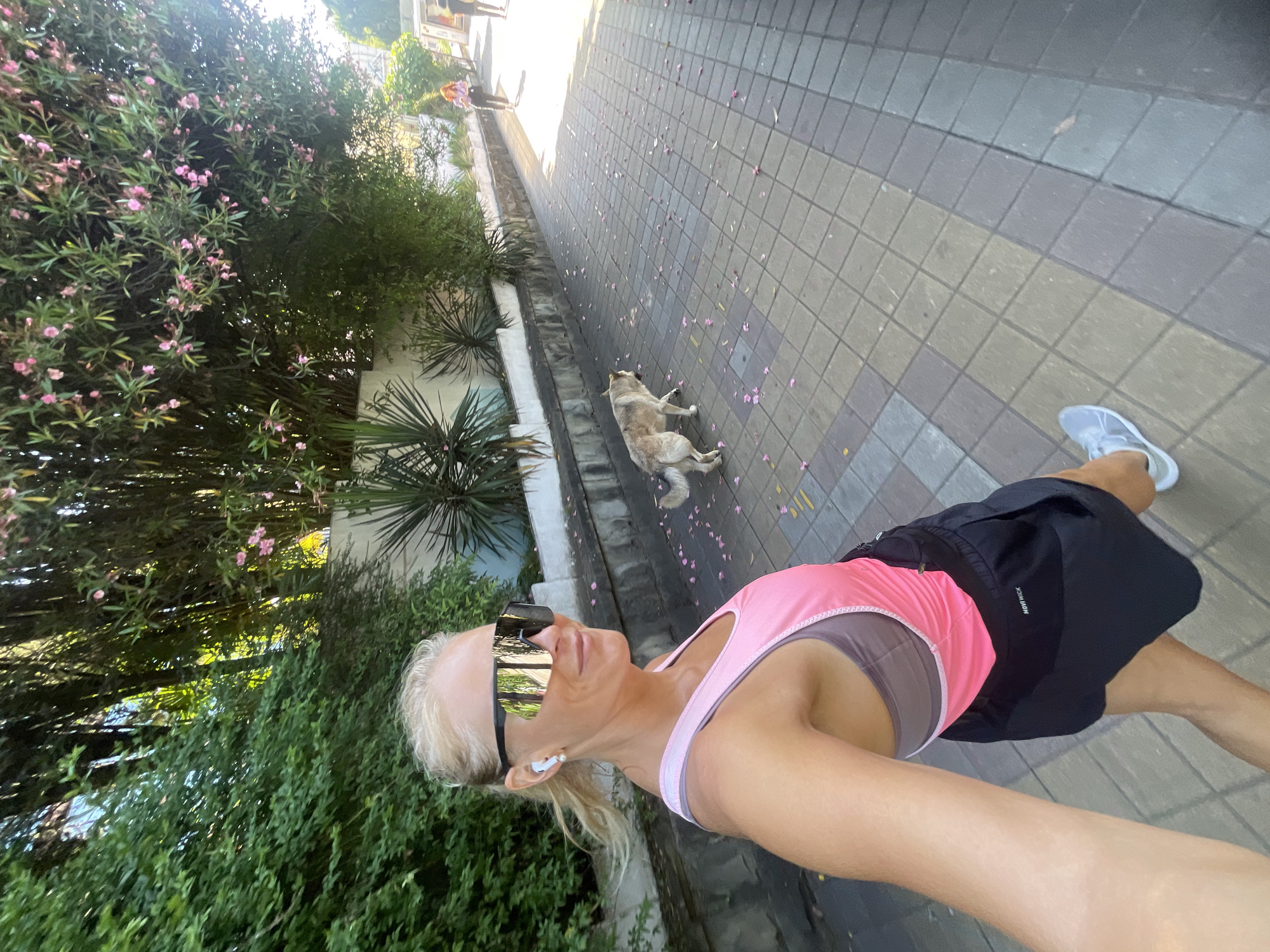 A blonde woman with large reflective sunglasses and white earbuds is taking a selfie while walking on a paved sidewalk. She is wearing a pink tank top and black athletic shorts. To her right, a medium-sized dog with tan and white fur walks away from the camera. The sidewalk is made of square tiles and is scattered with pink flower petals. Behind the woman is lush green foliage, including bamboo and other plants, with some pink flowers visible. A white wall runs along the edge of the foliage. In the background, other people are visible walking on the sidewalk. The scene appears to be outdoors on a sunny day, possibly in a park or along a promenade in Sochi, Russia, given the presence of sub-tropical vegetation and the paved walkway.
