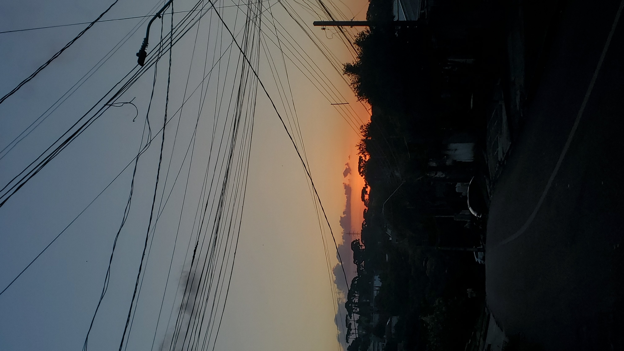 The image captures a residential street scene in Curitiba, Brazil, at either dusk or dawn. The sky transitions from a deep blue overhead to a vibrant orange and yellow hue along the horizon, where the sun is partially visible behind a silhouette of trees and scattered clouds. Numerous dark electrical and communication wires crisscross the upper half of the frame, with a street light fixture extending from the right and a loose wire segment visible.
The middle ground presents a dark, undulating landscape, heavily forested with tall, pointed trees, consistent with Paraná pines, and interspersed with dimly visible residential structures. A distant electrical transmission tower is faintly discernible on the left side of the horizon.
In the foreground, a paved asphalt road with a faded white centerline curves from the bottom center towards the left, disappearing into shadow. Two parked vehicles, one light-colored and one darker, are barely visible on the left side of the road. A utility pole stands on the right, supporting some of the overhead wires. The immediate foreground and sides of the road are obscured by deep shadows, revealing only indistinct outlines of vegetation and buildings. No individuals are present.