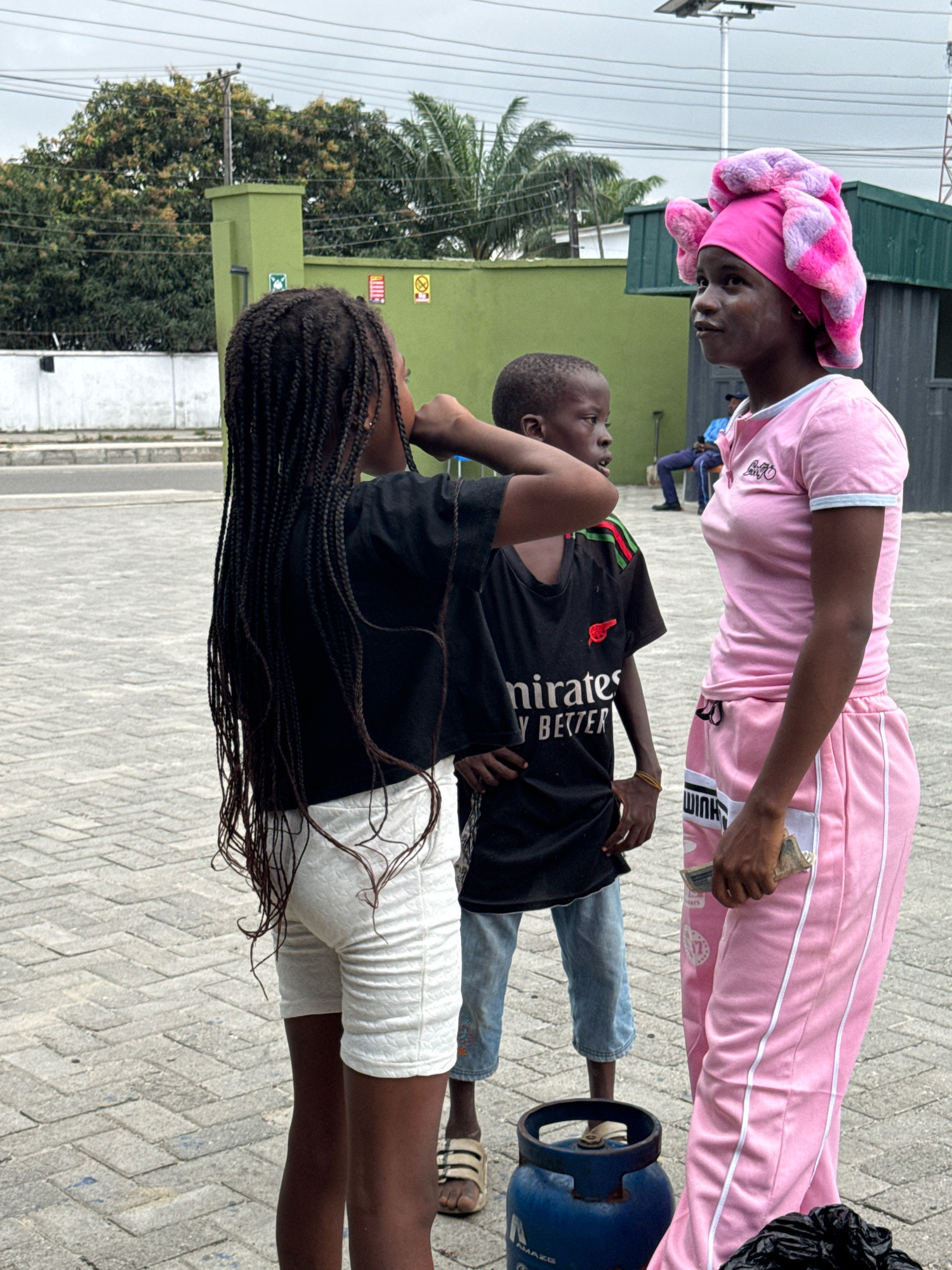 Three children are standing on a paved surface in what appears to be a commercial area in Ado, Nigeria. The child on the left, facing right, has long, dark braided hair and is wearing a black t-shirt and white textured shorts. The middle child, a boy facing left, wears a black t-shirt with "Emirates" and "Y BETTER" visible, along with light blue jeans. The child on the right, facing left, is wearing a pink t-shirt and pink tracksuit bottoms, and has a pink and purple textured head covering. A blue gas cylinder is visible in the foreground, to the left of the child on the right. In the background, there is a green building with trees and overhead power lines.