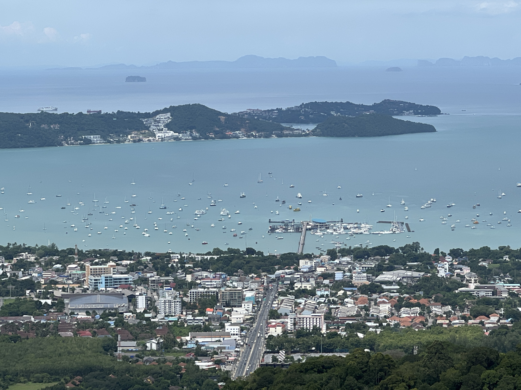 An aerial, wide-angle shot captures a panoramic view of Karon, Thailand, a coastal town nestled beside a vast expanse of turquoise ocean dotted with numerous sailboats and a few larger vessels. The cityscape is densely packed with buildings of varying sizes and architectural styles, ranging from low-rise residential homes with terracotta roofs to taller, modern hotels and commercial structures. A prominent road bisects the mid-ground of the city, with vehicles visible along its length. In the foreground, lush green trees and foliage create a natural border to the urban landscape. Beyond the city, a chain of verdant, hilly islands stretches towards the horizon, where distant mountains are faintly visible under a hazy sky. A pier with several moored boats extends into the ocean in the middle-ground, indicating a harbor or marina. The scene appears to be a daytime shot, with diffused light suggesting either an overcast sky or early morning/late afternoon conditions.