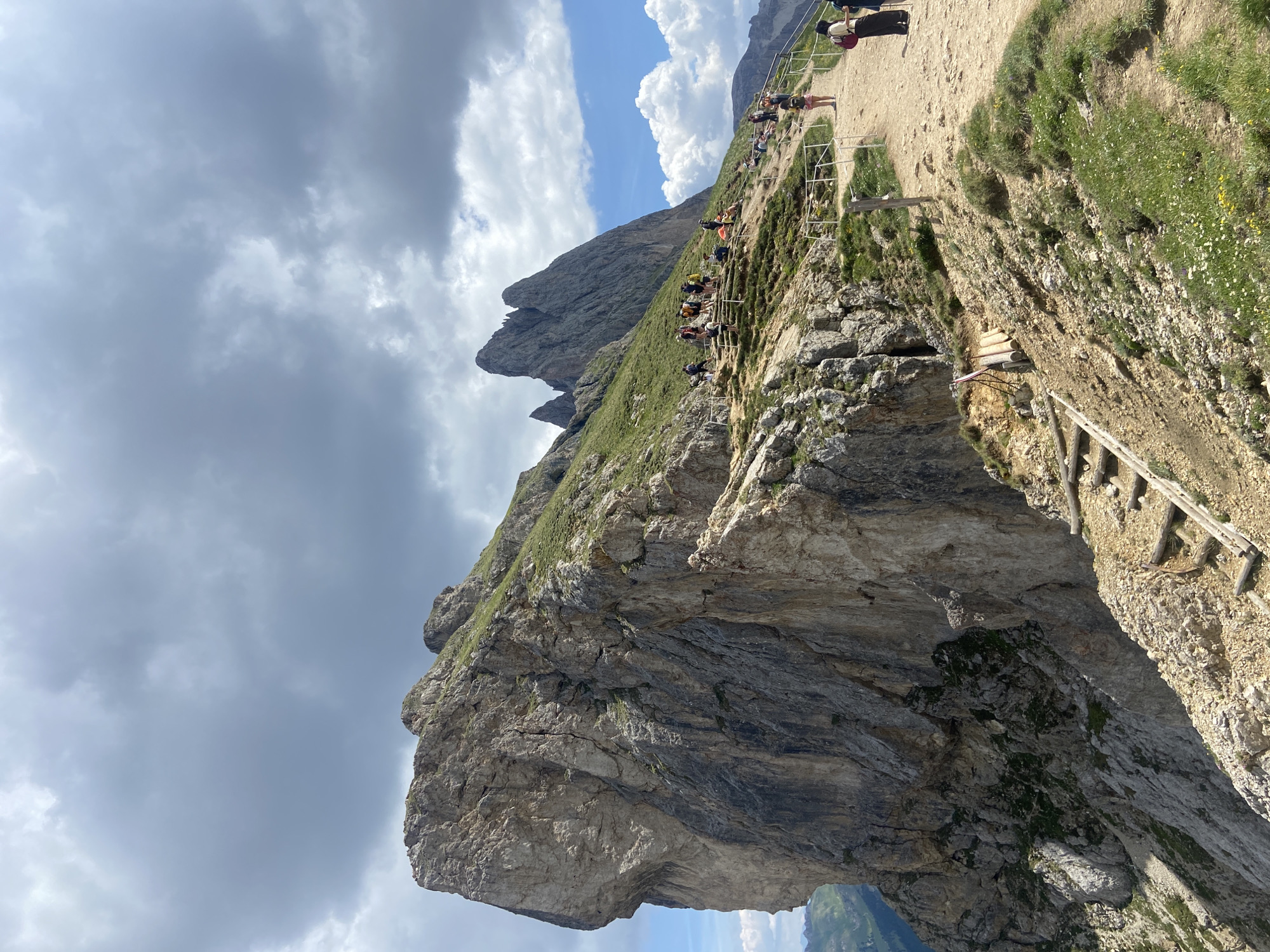 This media file captures a dramatic mountain landscape with a winding path and a group of people. The scene is characterized by steep, rocky cliffs covered in patches of green grass and wildflowers. A well-worn dirt path, bordered by a safety railing, snakes along the mountainside, indicating it is a popular hiking route. Several individuals are visible on the path, some walking, others pausing to admire the view.
The sky is filled with a mix of dramatic, dark clouds and patches of bright blue, suggesting a dynamic and possibly changing weather pattern. The time of day appears to be daytime, likely late morning or early afternoon, given the bright lighting and the shadows cast by the rocks and people.
A prominent geological feature is a large, overhanging rock formation that juts out over a steep drop, creating a sense of awe and perhaps a touch of trepidation. Wooden steps are built into the side of the mountain in some areas, providing access and support for hikers. The overall impression is one of a challenging yet rewarding outdoor adventure in a rugged, natural environment. There is no visible text in the image.