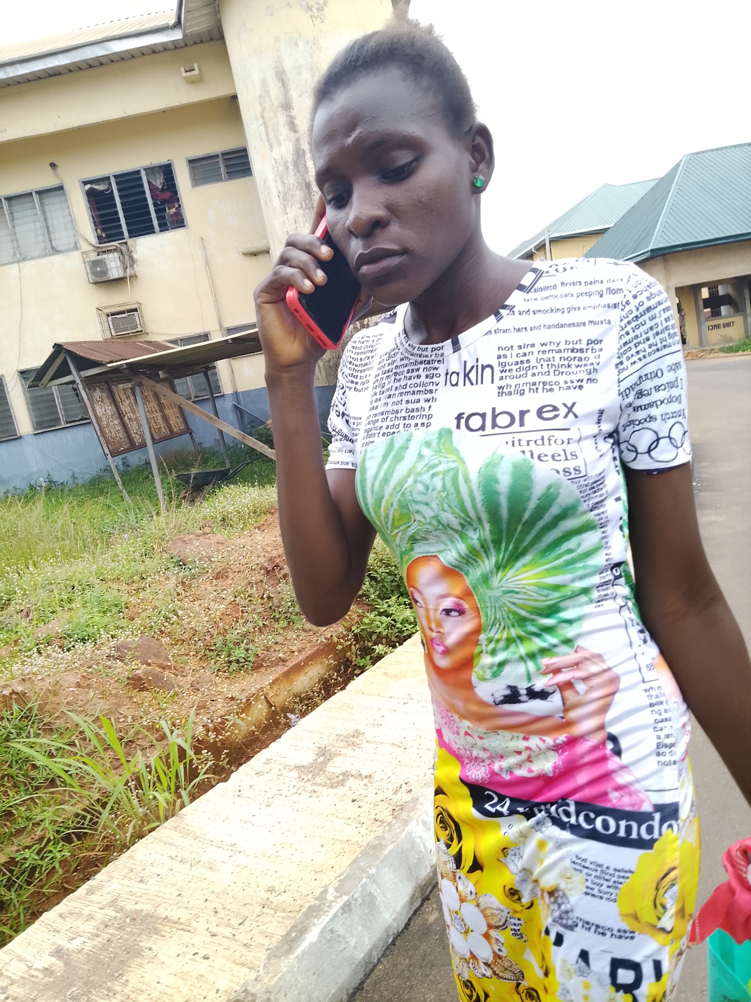 In the city of Nsugbe, Nigeria, a young woman, viewed from the chest down, is captured mid-conversation while holding a red smartphone to her right ear. She is wearing a vibrant, white t-shirt adorned with a collage of text and images. Prominently displayed on the shirt is a graphic of a woman's face with green, leafy hair and a colorful, floral pattern on the lower portion, including yellow roses and the text "24dcond". The woman stands outdoors, with a light-colored concrete barrier in the foreground and overgrown vegetation to her left. In the background, a pale yellow building with multiple windows and an air conditioning unit is visible, along with a covered structure with a tin roof. The overall setting appears to be a casual outdoor environment, possibly near an educational institution or residential area.
