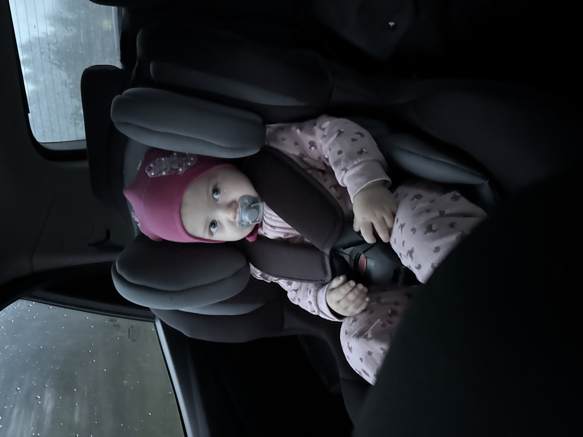 A baby is sitting in a car seat. The baby is wearing a pink hat and pajamas. A pacifier is in the baby's mouth. The car window is visible and shows rain droplets. The photo was taken inside a car in Сосново, Russia.