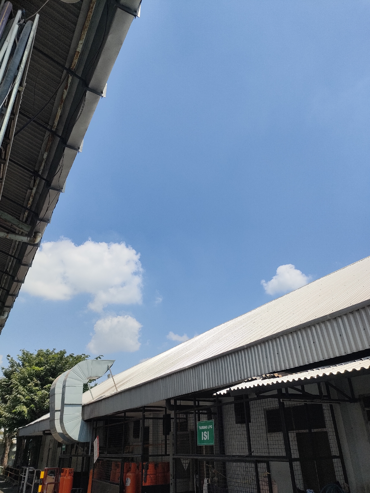 An exterior medium shot captures a bright, sunny day in Sayung, Indonesia, with a clear blue sky dominating the upper portion of the frame. The image is divided by the sloping corrugated metal roofs of two buildings, one on the left extending from the top edge and another on the right occupying the lower half.
On the left, a dark, weathered roof with visible metal supports and a few wires hangs over the scene. This roof partially obscures the view of a large, grey, curved ventilation duct that emerges from the building on the right and extends upwards.
The main structure in the lower right is a long, low-rise building with a white, ribbed metal roof. Below the roofline, a section of the building is visible, featuring a caged enclosure with dark metal bars. Inside the enclosure, obscured by shadows and wire mesh, are dark rectangular shapes that could be equipment or stored items. A green sign with white text, reading "TABUNG LPG ISI," is attached to the front of this enclosure.
To the left of this building, a lush green tree with dense foliage provides a stark contrast to the industrial structures. In the foreground, partially visible at the bottom left and bottom right of the frame, are several orange LPG cylinders, some upright and others lying on their sides, indicating a potential storage or distribution area. The ground appears to be paved or made of dark material. The overall impression is of an industrial or commercial outdoor space.
