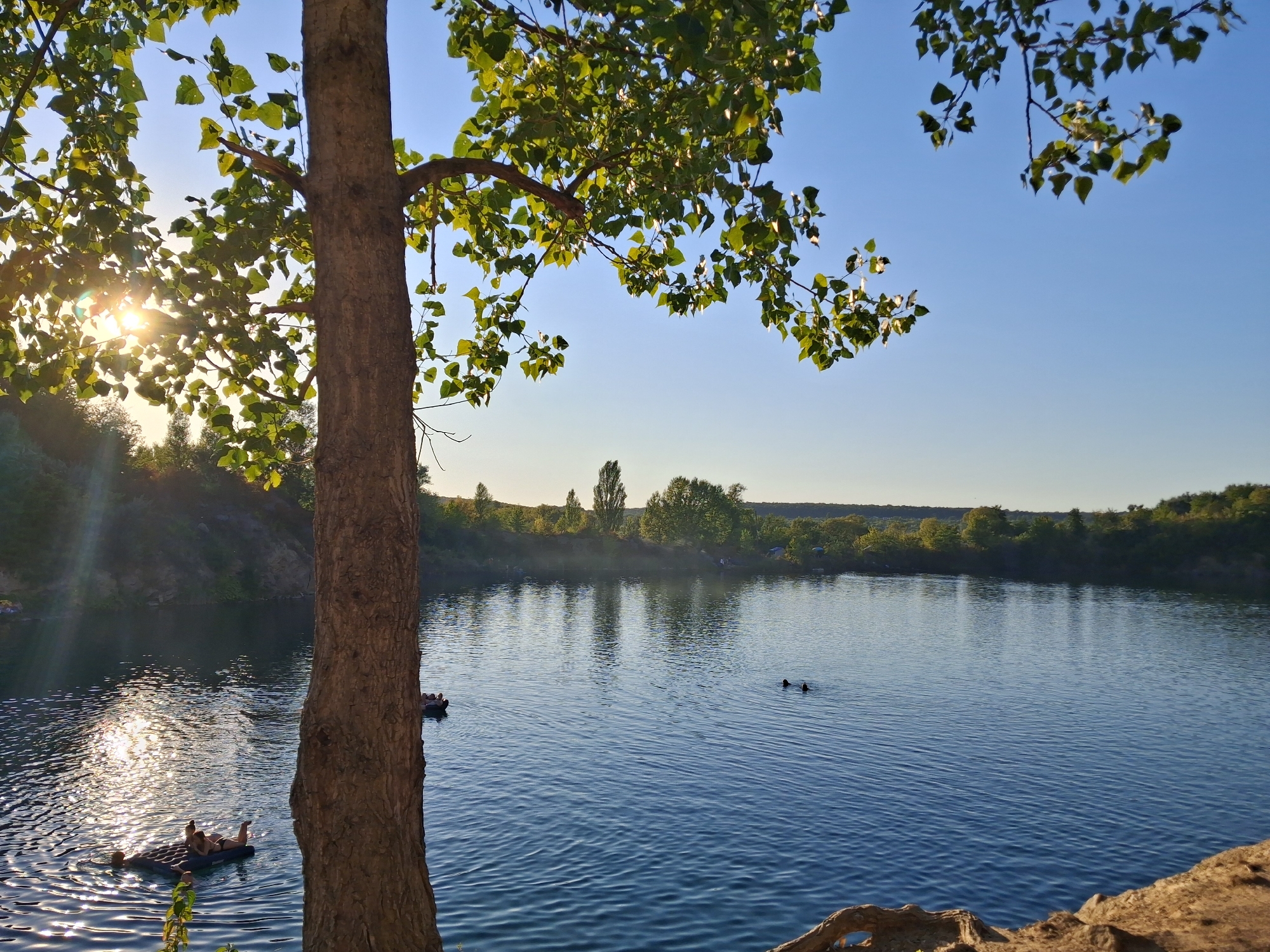 The image depicts a tranquil scene at the Kashperivka quarry in Ukraine, bathed in the warm glow of the setting sun. A large tree trunk dominates the foreground, its rough bark contrasting with the smooth, rippling surface of the quarry lake. Sunlight glints off the water, creating a shimmering effect.
Several individuals are enjoying the water. In the left foreground, two people are relaxing on a large, inflatable mattress. Further out in the lake, two more figures are visible, seemingly swimming or wading. On the right side of the lake, a person is seen on another inflatable tube.
The quarry's rocky banks rise gently on the far side of the lake, covered in lush green vegetation and trees. The sky above is a clear, serene blue, with a few branches from the foreground tree reaching into the frame, their leaves catching the sunlight. The overall impression is one of peaceful leisure and natural beauty.
