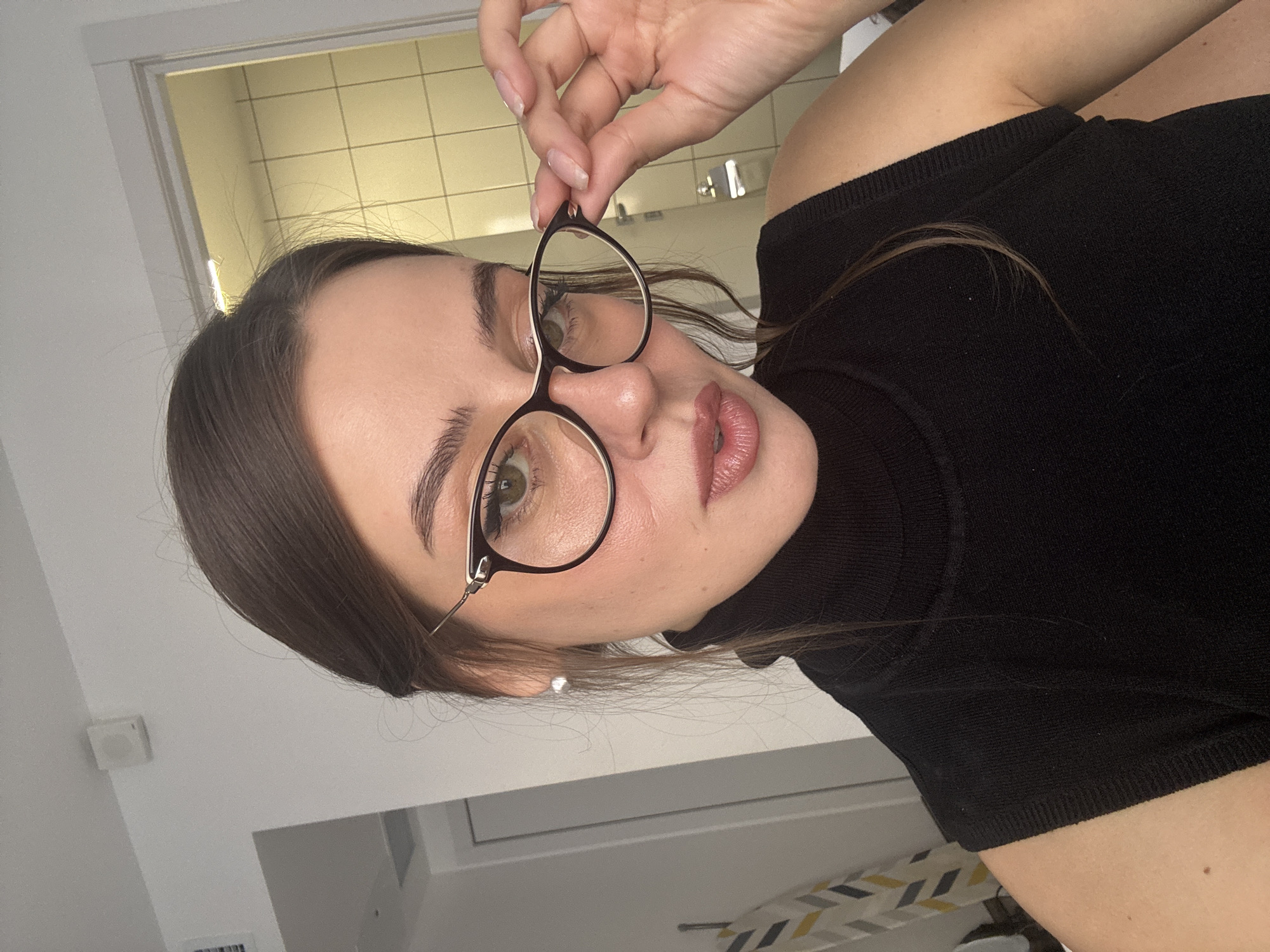 A woman with light olive skin, light brown hair, and light green eyes holds eyeglasses. She wears a black turtleneck shirt. The background includes a white wall, a door frame, and a tiled wall possibly in a bathroom. The location is not discernible as Moscow, Russia, as no identifiable landmarks or cues indicating the city are visible.
