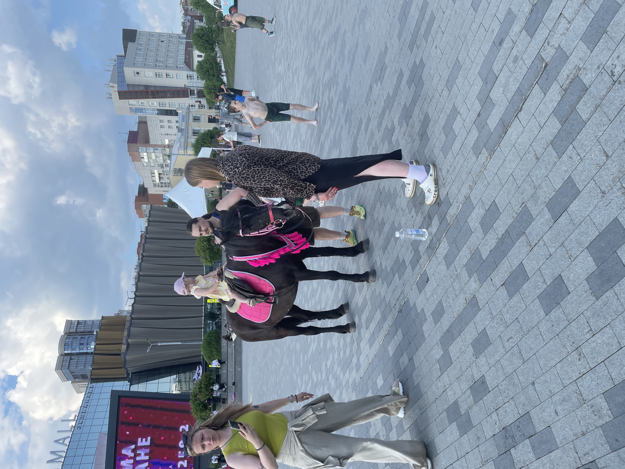 The image depicts a street scene in Perm, Russia, with a focus on a horse being held by two individuals. The setting is an outdoor area paved with grey bricks, against a backdrop of modern buildings, including a structure labeled "MALL". One individual is dressed in a leopard-print jacket and a black skirt, while the other individual holds the horse's reins. A young child wearing a pink cap sits on the horse's back. A discarded water bottle lies on the ground near the group. In the background, several shirtless men are visible, and another woman stands closer to the foreground, using a mobile phone. The sky is partly cloudy.