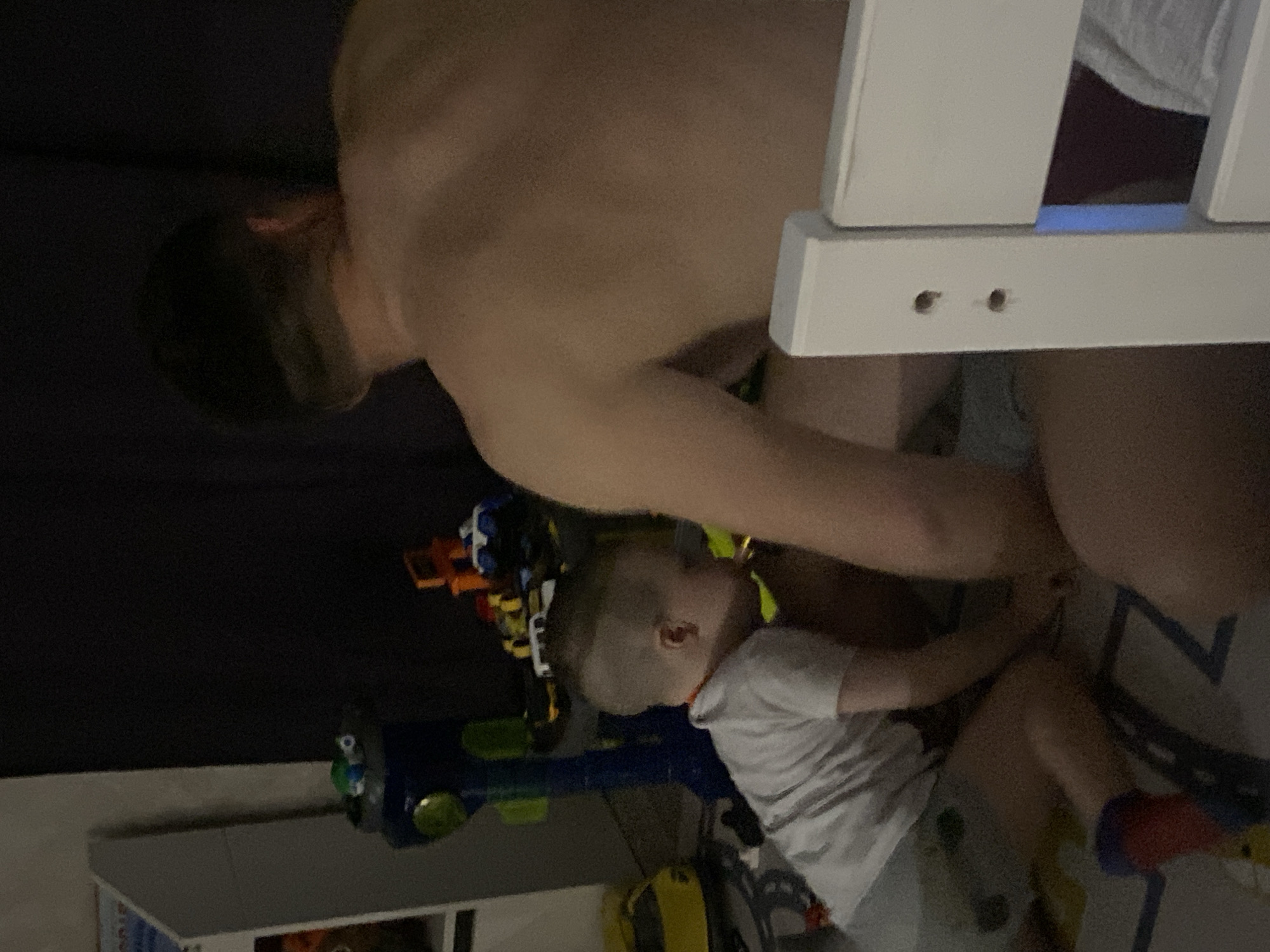 A shirtless adult is near a child who is on the floor surrounded by toys. The location is unknown, but it is not in the city of Sochi, Russia.