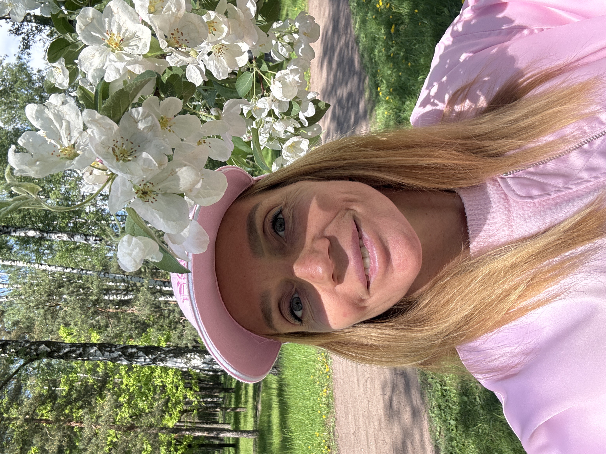 A woman with blonde hair and blue eyes is wearing a pink hat and a pink jacket. She is smiling and looking at the camera. To her left is a tree branch with white flowers. Behind her is a park with green grass and trees. A path is visible. This image appears to be taken in a park, likely within Saint Petersburg, Russia, due to its common architectural style of trees.