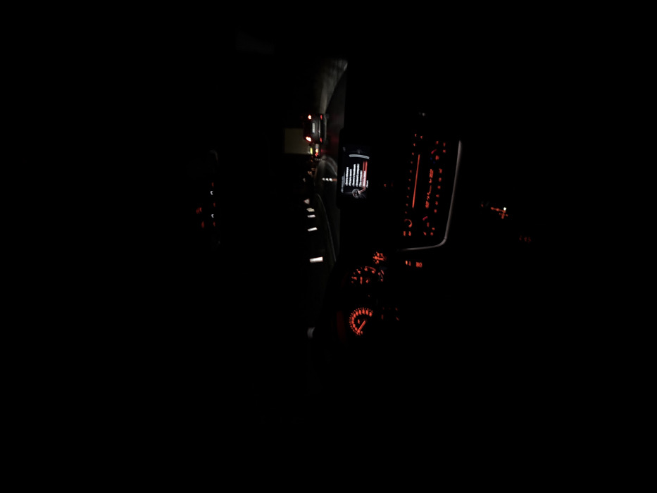 The image depicts the interior of a vehicle at night, viewed from the driver's perspective on a road in Poleto, Bulgaria. The dashboard is illuminated with red lighting, displaying a speedometer on the left and a tachometer on the right. A central infotainment screen shows a menu or text, and below it, climate control buttons are also illuminated in red.
Through the windshield, a dark, wet-appearing road is visible. White reflective road markers line the left side of the thoroughfare, curving slightly to the right. Ahead, multiple vehicles are present, their red taillights distinctly visible against the darkness, indicating motion or a stopped queue. One vehicle, a dark-colored passenger car, is directly in front, while another larger vehicle, possibly a truck, is further ahead on the right. No individuals are discernible within the vehicle or on the road.