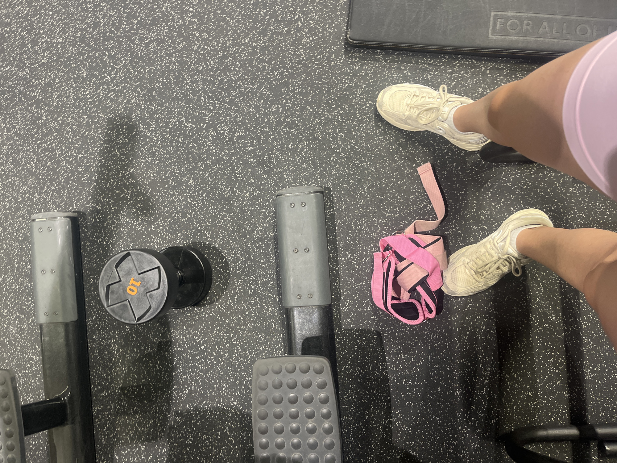 The image shows a person at a gym in Saarbrücken, Germany. The person is wearing white sneakers and light purple athletic shorts. They are standing on a dark gray rubber floor. To their left is a piece of gym equipment with a 10 kg dumbbell attached to it. Next to their foot is a pink and black weightlifting belt.