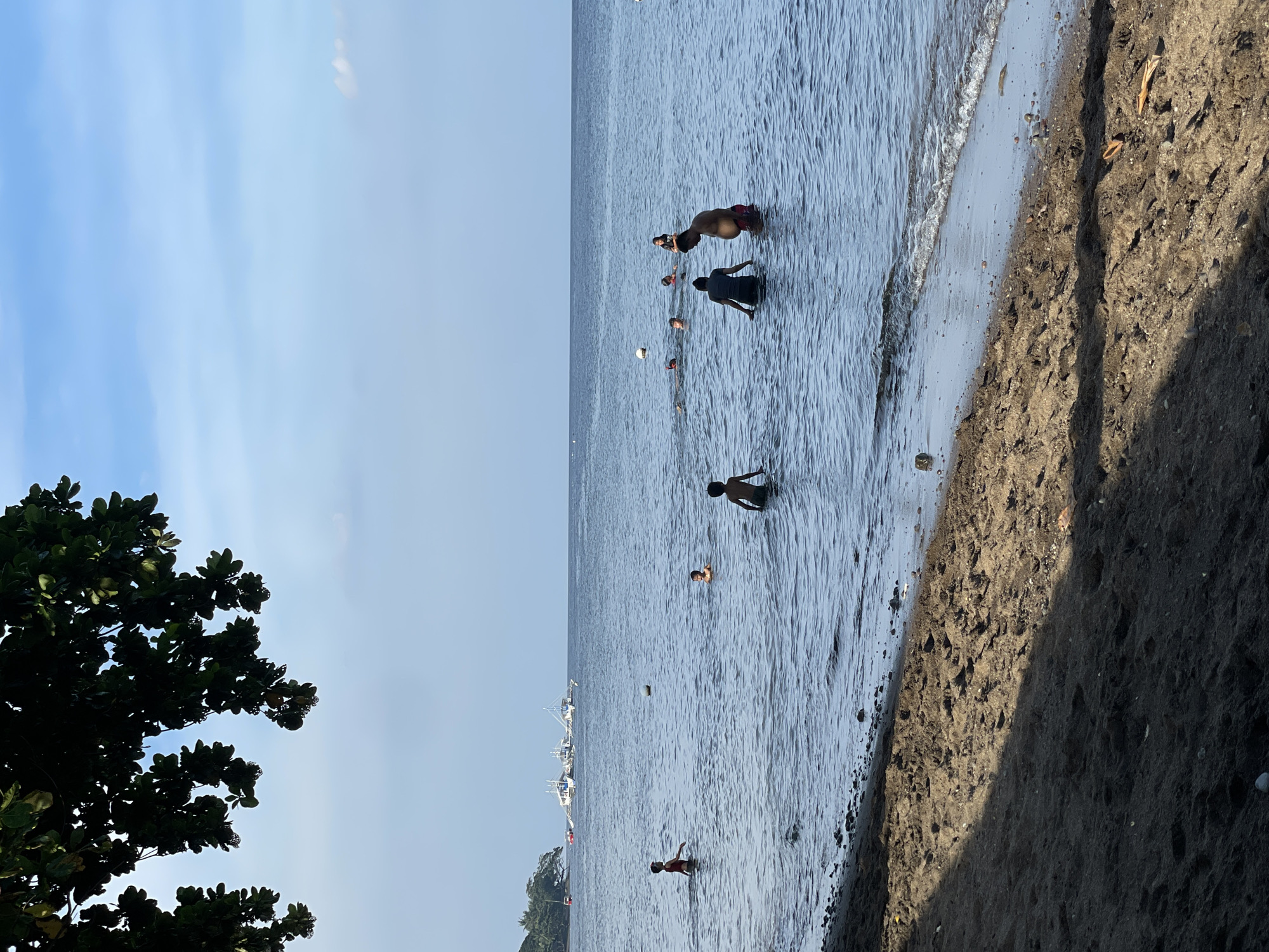 The image depicts a tropical beach scene in Dauin, Philippines. In the foreground, dark sand is visible, with a shadow cast across it from an overhead tree. The water is calm with gentle waves lapping the shore. Several people, appearing to be of various ages, are in the water, some wading and others swimming. In the mid-ground, a white ball floats on the surface. Further out in the ocean, several white boats are anchored, and a treeline is visible on a distant shore. The sky is a clear blue with some scattered white clouds.