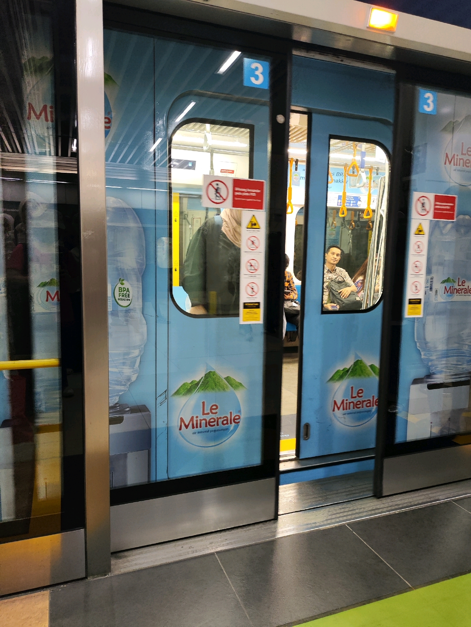 The image captures the exterior of a train carriage with its doors open at a station platform. The train doors are adorned with large graphics of Le Minerale bottled water, featuring green mountains and the brand's logo. Several safety signs are visible on the doors, including a prohibition sign for smoking and a warning sign with a yellow triangle. Inside the train, passengers can be seen through the glass, some standing and others seated. One man is clearly visible inside, looking towards the camera. The platform appears to be made of dark tiles, with a bright green section visible in the foreground. The overall scene suggests an urban transportation hub in Central Jakarta, Indonesia, likely a train station.