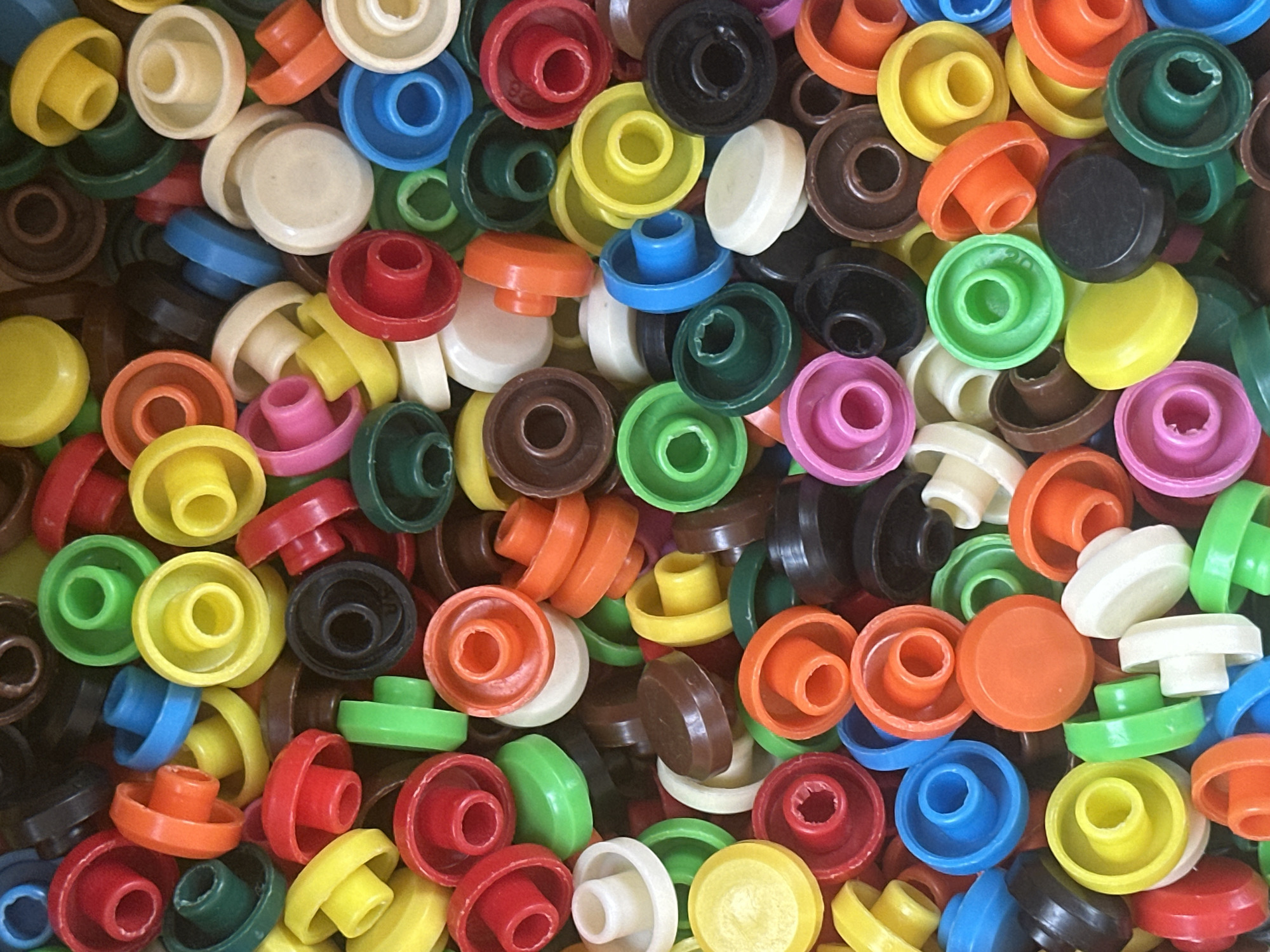 The image displays a chaotic, overhead view of a large collection of colorful, plastic toy pieces. The objects are predominantly circular or disc-shaped, with various textures and raised elements, suggesting they are building or stacking components from a children's toy set. The colors are vibrant, including red, blue, green, yellow, orange, pink, black, brown, and white. The pieces are piled and jumbled together, with no discernible order or arrangement. Some pieces appear to be upside down or partially obscured by others. The lighting is even, indicating an indoor setting, and there are no shadows that suggest a specific time of day. The overall impression is one of a toy box or a scattered assortment of playthings. No people, text, or specific location cues are visible.
