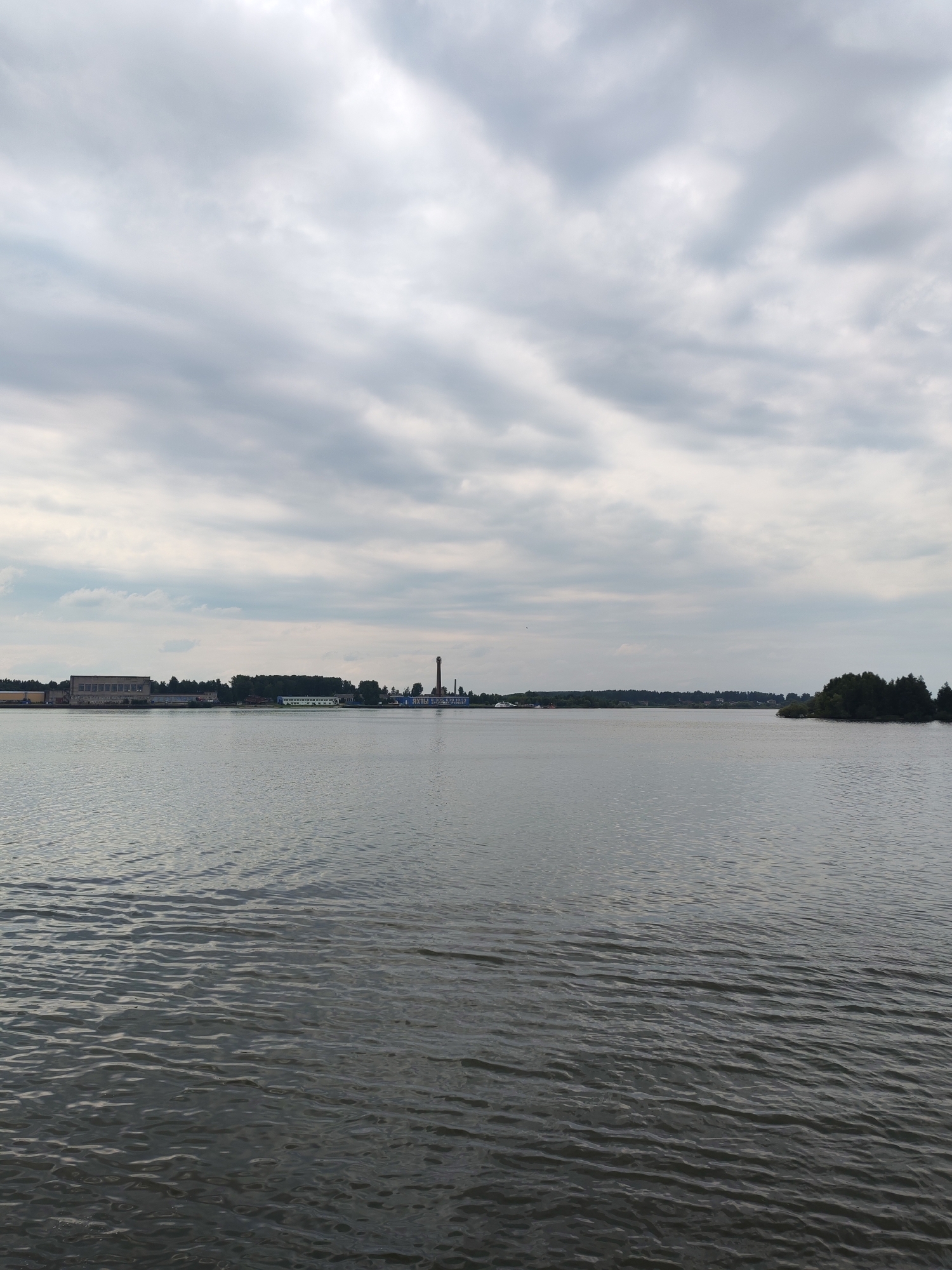 This is a photograph taken from a body of water, likely a river or lake, looking towards a distant shoreline. The sky is overcast with a thick layer of grey clouds. The water in the foreground is dark and shows gentle ripples.
The shoreline in the middle ground features a line of trees and several industrial buildings. Prominently visible are a long, low-slung building with a flat roof on the left, a taller brick chimney with a thin plume of smoke, and other structures with blue and green accents. The overall impression is of an industrial area bordering a natural body of water. The location is identified as Bol'shie Mkhi, Russia. There are no people visible in the image.