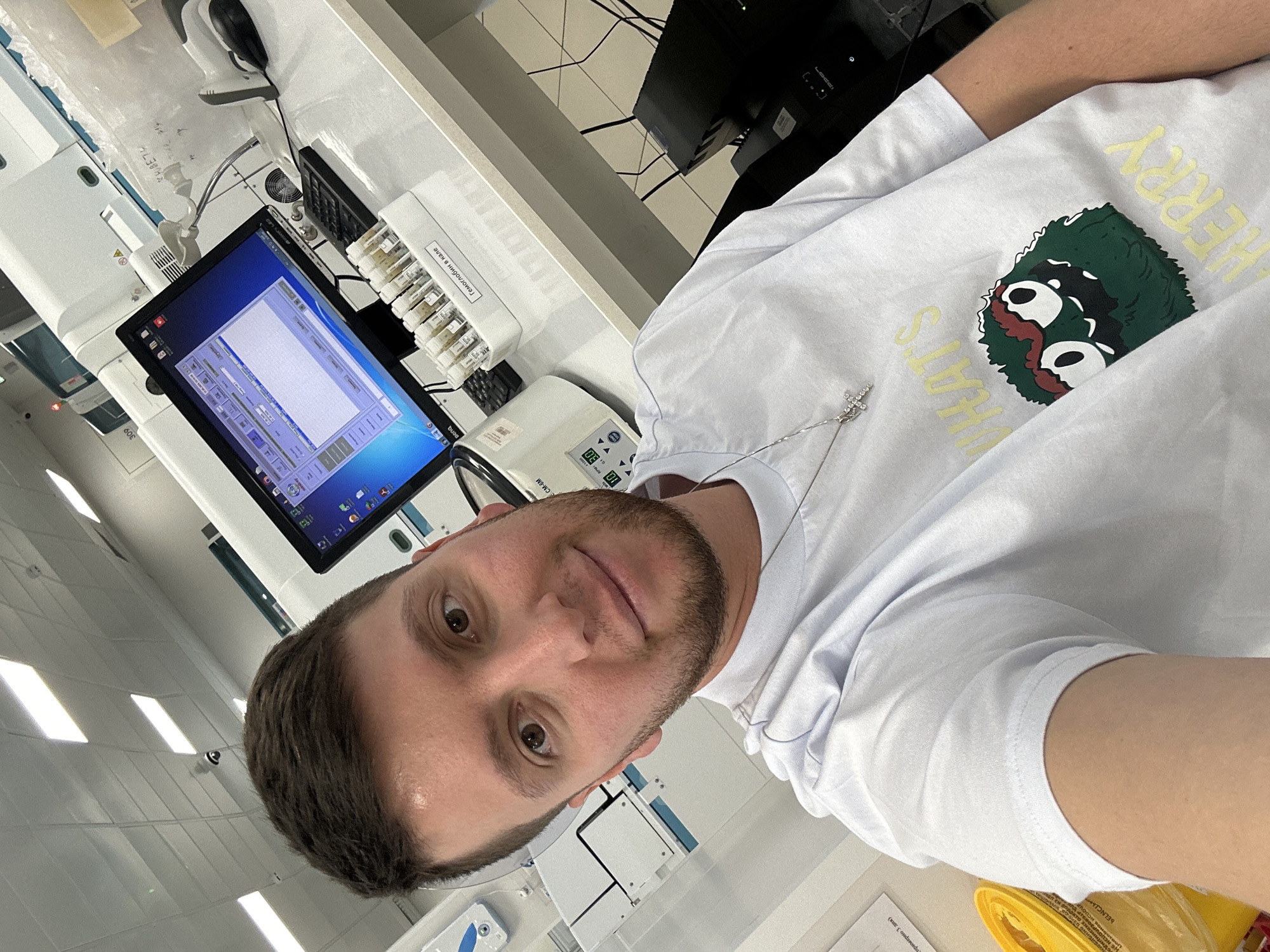 A man wearing a white t-shirt with a cartoon graphic and a cross pendant is taking a selfie indoors. The room appears to be a laboratory or medical facility, with diagnostic equipment and vials of samples visible on a counter next to a computer monitor displaying data. Text and labels are visible on the equipment, including the phrase "Гемоглобин в кале" (Hemoglobin in feces) written in Russian. The environment suggests a clinical or scientific setting. Moscow, Russia cannot be confirmed as the location based on the image alone.