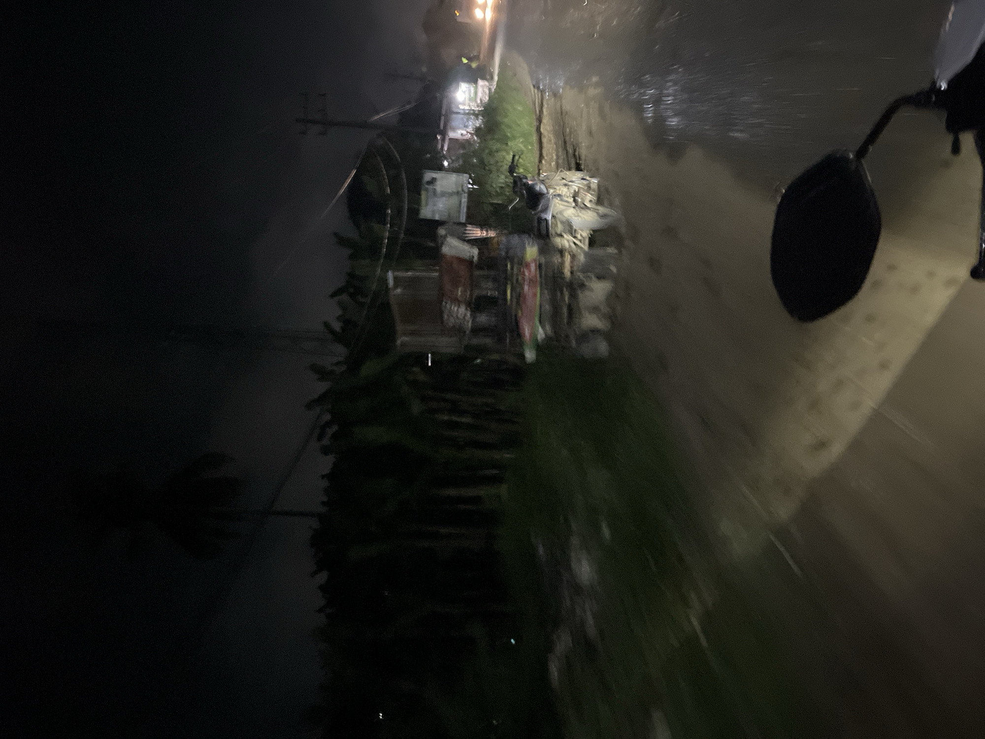 The image depicts a dark, nighttime scene, likely taken from a moving vehicle, possibly a motorcycle, as indicated by the side mirror in the foreground. The overall impression is one of a rural or semi-rural area in Indonesia, specifically Benuaraja.
The left side of the image is dominated by deep shadow, with a hint of a palm tree silhouette visible. A utility pole with wires stretches upwards, suggesting the presence of infrastructure. In the center and right of the frame, the scene is more illuminated, revealing a roadside environment. There are several makeshift structures, appearing to be small stalls or vendors, with some goods displayed. These structures are partially obscured by foliage and debris. The ground appears wet and reflective, indicating recent rain or a damp atmosphere, consistent with a tropical climate at night.
The motion blur in the image suggests the viewer is in transit, moving along a path or road. The lighting is dim and artificial, with a few indistinct lights visible in the distance, possibly from buildings or other vehicles. The atmosphere feels a bit desolate due to the darkness and the low visibility. No people are clearly discernible, though the presence of the structures implies human activity. The focus is primarily on the environment and the general atmosphere of a wet, dimly lit Indonesian night. There is no visible text in the image.