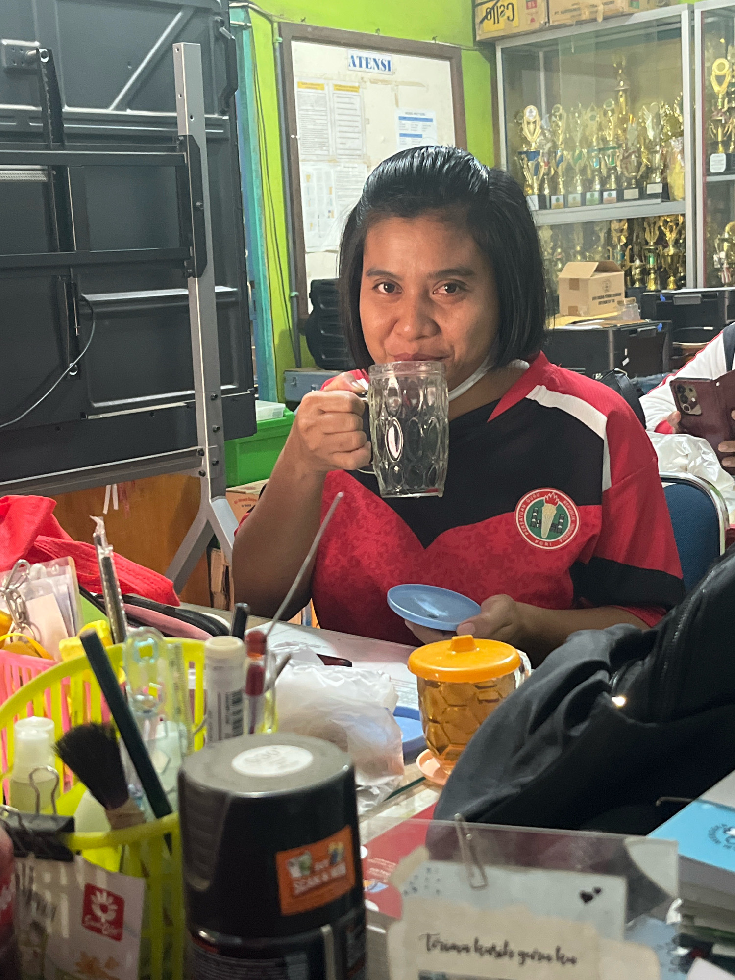 A medium shot captures a woman with dark hair and brown eyes in the foreground, seated at a table and holding a clear glass mug to her lips. She is wearing a red and black shirt with a circular emblem on the left chest. In front of her, on the table, are various objects including a blue saucer, an orange lidded container, and a black bag. To the left, a bright yellow basket contains a collection of items such as pens, a small brush, and bottles. The background reveals a cluttered environment with shelves filled with trophies, a bulletin board with papers, and what appears to be electronic equipment. The overall setting suggests an indoor space, possibly an office or a common area, in Meliau Hulu, Indonesia.