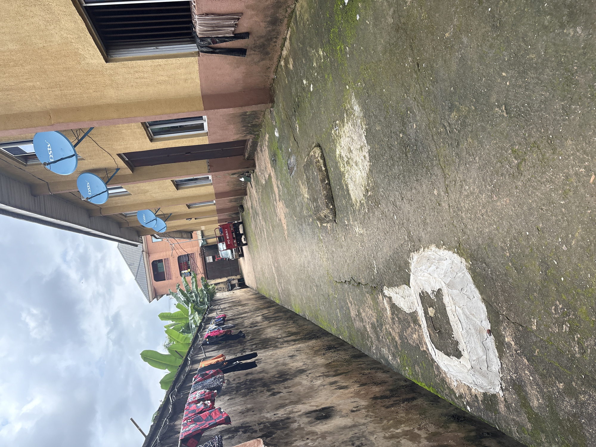 This is a low-angle, eye-level shot of a narrow concrete alleyway in Onitsha, Nigeria, lined by buildings and a textured wall. The wall on the left, covered in grime and moss, has laundry hanging on a clothesline. Banana trees are visible above the wall. The building on the right is yellow with pink trim and has several satellite dishes mounted on its facade. Clothes are also hanging outside of a window on this side. A small red three-wheeled vehicle is parked in the alley, and in the distance, another building with a metal roof is visible. The ground is cracked and mossy, with a patch of white paint or plaster in the foreground. The sky is overcast.