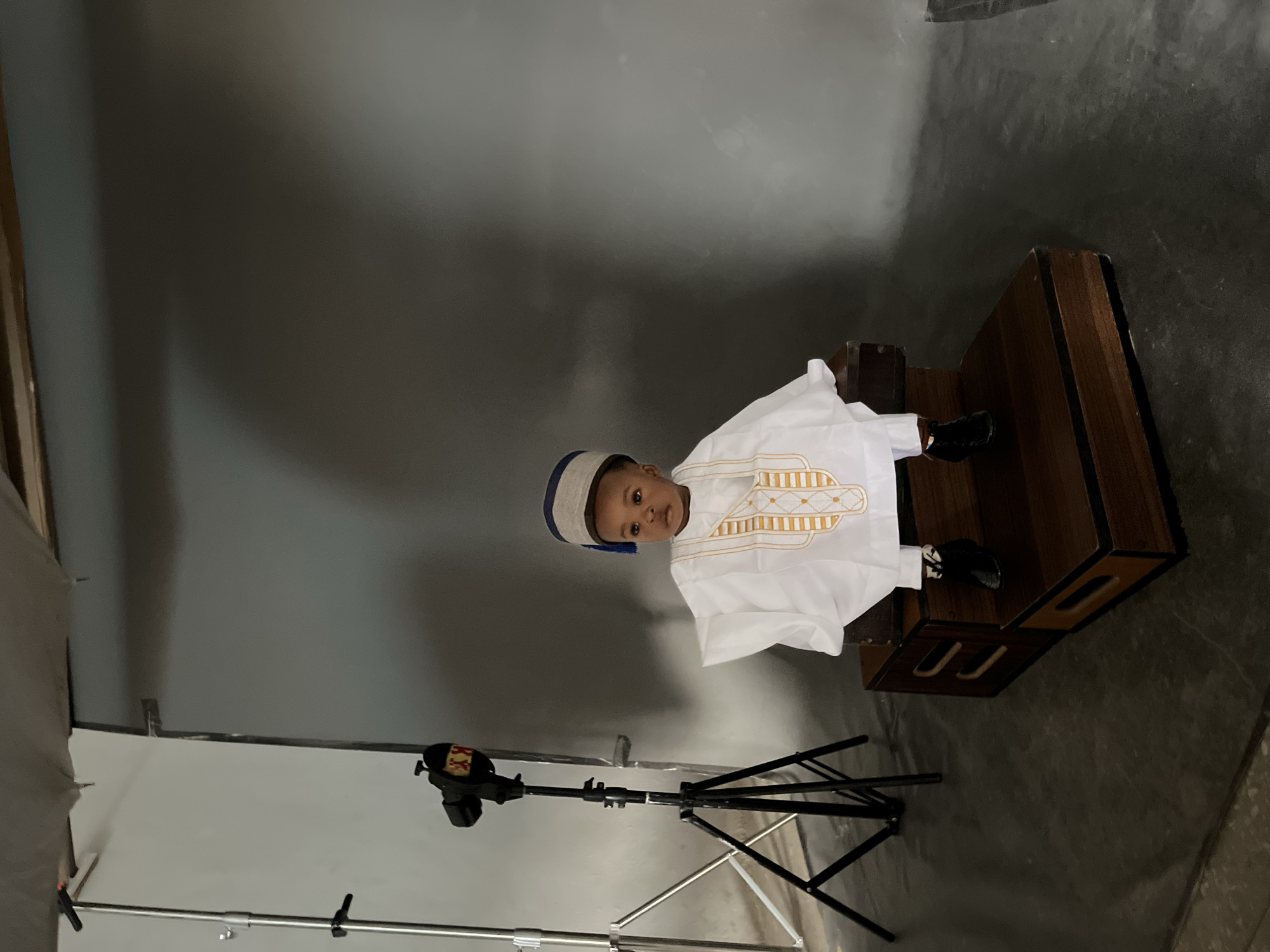 A young child, dressed in a white traditional Nigerian outfit and a matching hat, sits on a wooden platform in what appears to be a photography studio. The child is facing forward and looking towards the camera. The background is a gray studio backdrop. To the left of the child, a lighting stand with a studio light is visible. The flooring is a dark gray concrete. The scene is well-lit, indicating a professional photography setup. The child's attire suggests a cultural or celebratory occasion. The overall atmosphere is serene and posed, typical of a portrait session. No specific landmarks in Gura Top, Nigeria are discernible.