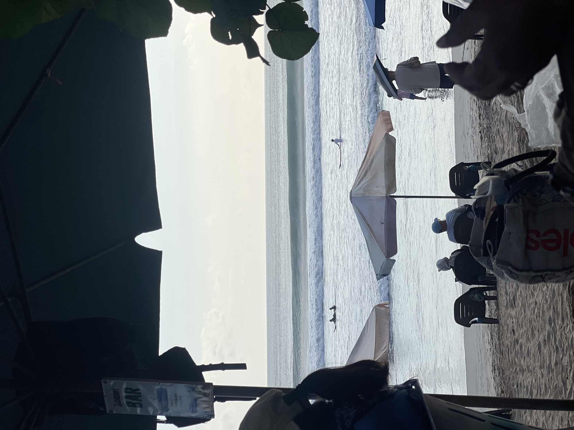 In Kuta, Indonesia, on a sandy beach, a view is captured from beneath a dark canopy, possibly an umbrella or awning. A sign for "CHERRY BAR" is visible on a pole to the left. In the ocean, several surfers are spotted on their boards, with waves breaking in the background under an overcast sky. The foreground features beach chairs and umbrellas, with some people seated, facing the water. One person stands in the shallow water holding an open umbrella. The scene depicts a relaxed beach setting.