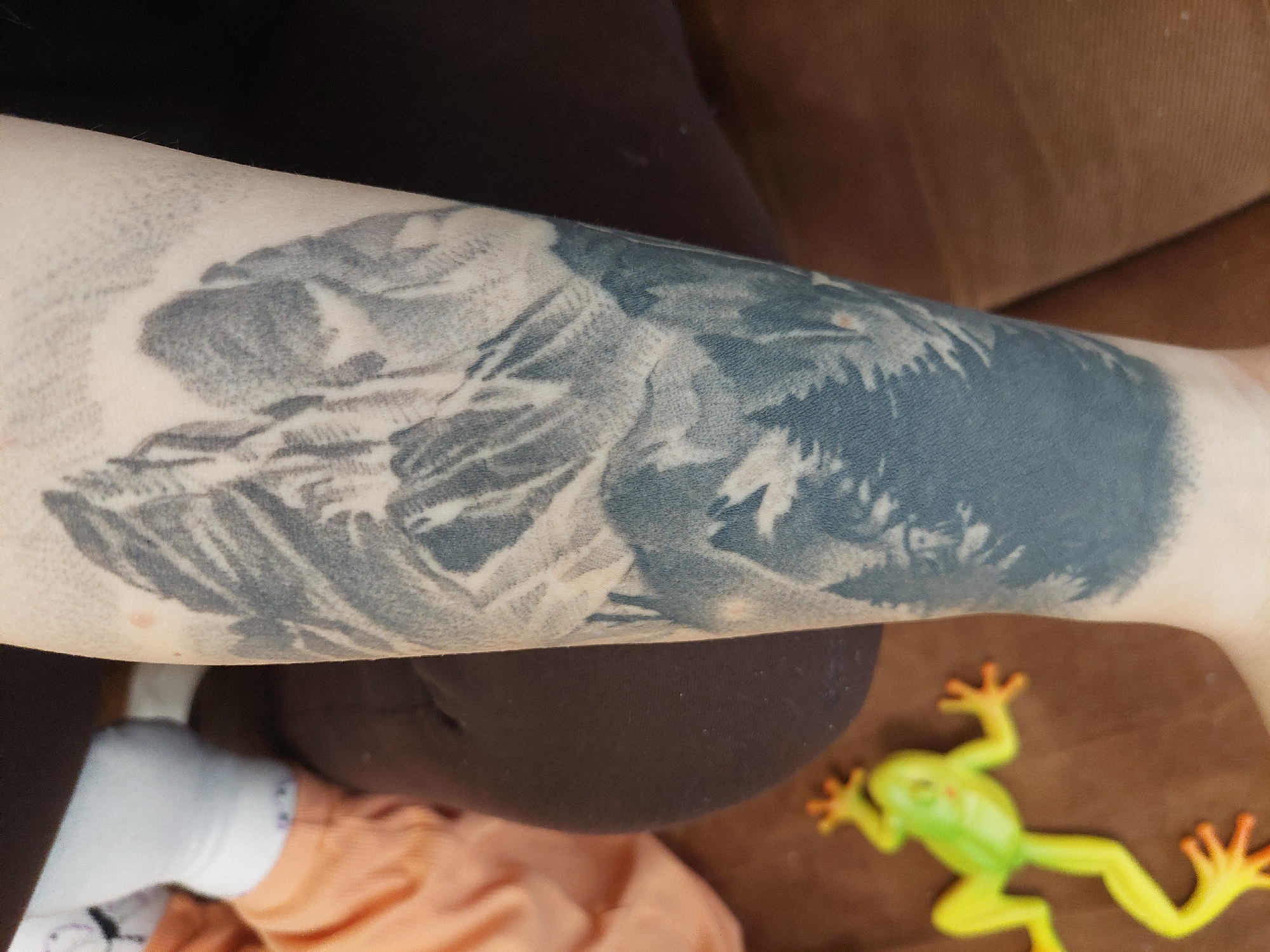 An arm with a grey tattoo, partially obscured by dark clothing, rests on brown fabric. A green plastic frog with orange details lies near the arm. A child's foot in a white sock and peach-colored clothing is visible in the lower left corner. The location in Plovdiv, Bulgaria is unknown.