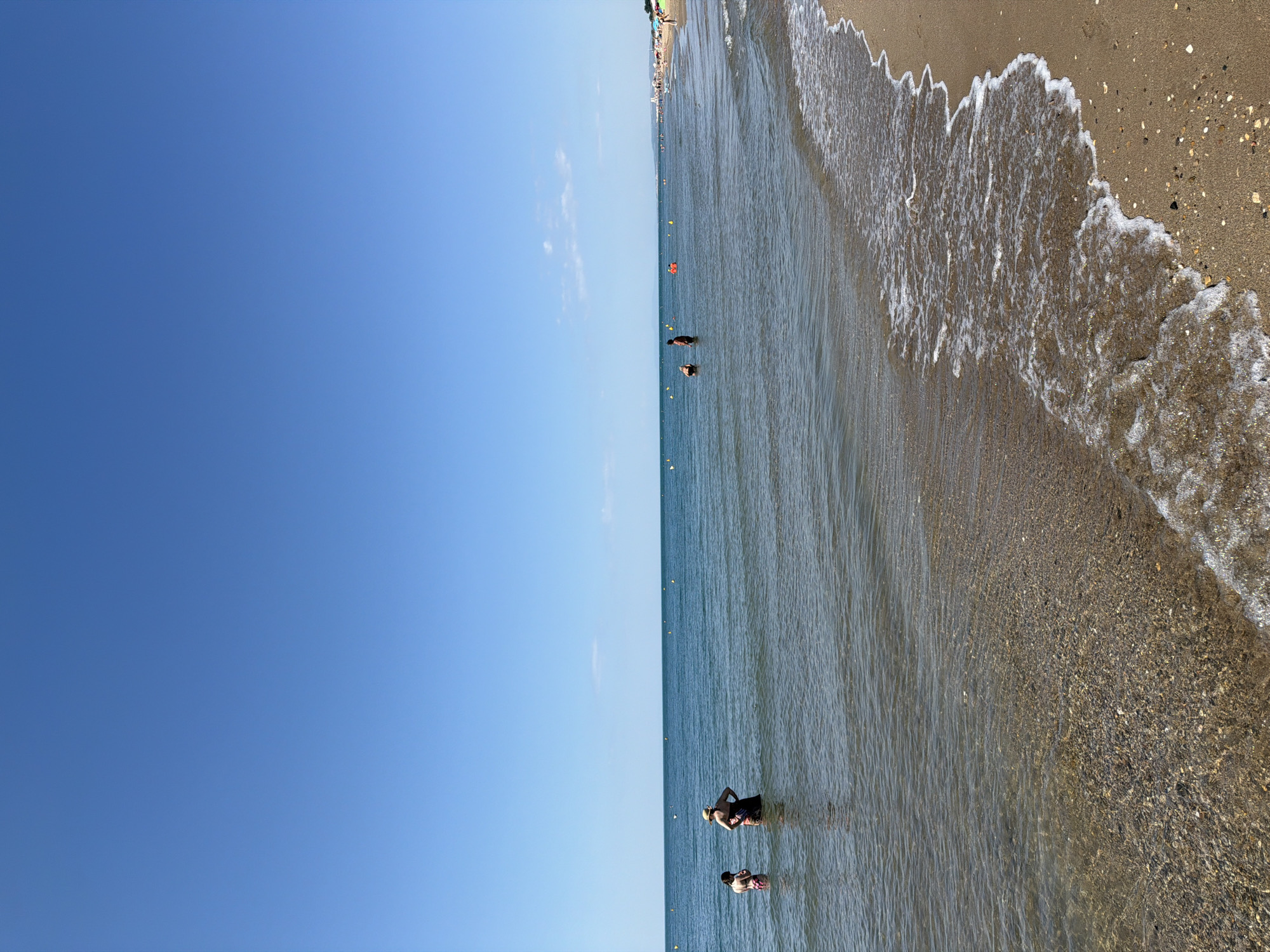 The image depicts a wide view of a sandy beach and the calm Mediterranean Sea under a clear blue sky. Several people are visible in the shallow water, some wading and others standing. On the left side of the frame, two individuals are in the water, one appearing to be an adult wearing a hat and a patterned wrap, and another a smaller figure. Further out, more people are scattered across the water, and some are visible on the distant shoreline, suggesting a populated beach. Yellow buoys are dotted in the sea, marking an area. The foreground shows the wet sand meeting the gentle waves, with small shells and pebbles visible on the beach. The scene captures a peaceful, summery day at the coast. This beach is likely Plage de la Grande Maïre or Plage de la Tamarissière in Sérignan, France, known for its long sandy stretches.