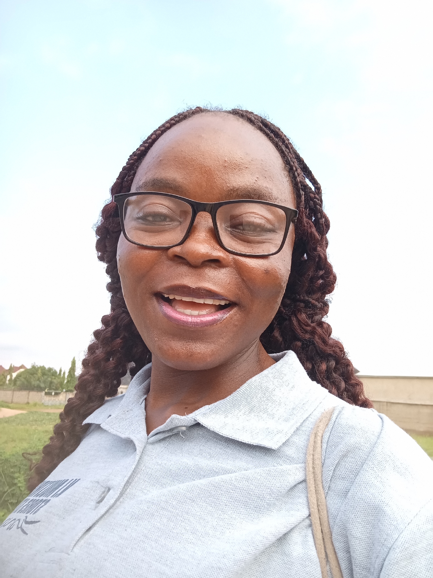 A young African woman wearing glasses and a grey polo shirt smiles at the camera. Her hair is braided with dark brown extensions. She is standing outdoors with a light blue sky in the background. In the distance, there are trees and buildings. The overall scene suggests a casual, outdoor setting in Mayo Dasa, Nigeria.