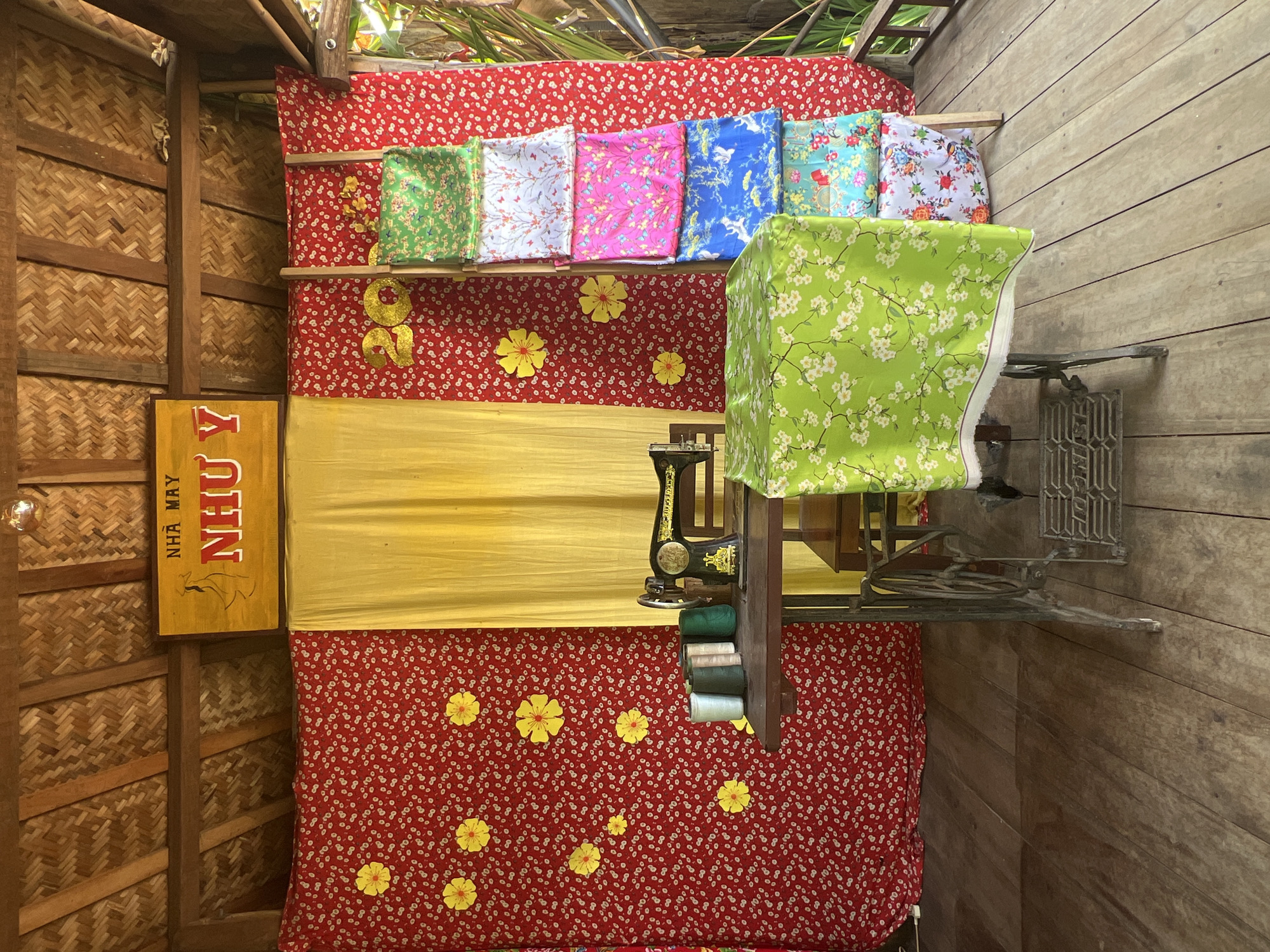This image depicts an outdoor stall in Hoi An, Vietnam, likely a tailor shop or a fabric vendor. The stall is set up on a wooden deck, with a thatched roof overhead. The background is dominated by red fabric with a white floral pattern and yellow accents, along with a yellow curtain in the center. To the right, several bolts of colorful patterned fabric are displayed vertically on a wooden ladder. In the foreground, a vintage sewing machine sits on a wooden table, surrounded by spools of thread. A large piece of green fabric with a floral print is draped over the sewing machine and table. A yellow sign with Vietnamese text and a silhouette of a woman hangs on the wall. The overall impression is one of a traditional craft workshop or market stall.