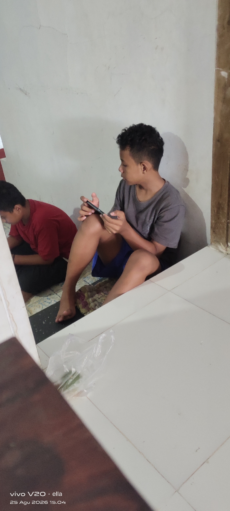Two young males are seated on the floor indoors, near a wall and a tiled surface. The male on the right, with short, curly dark hair, wears a gray t-shirt and blue shorts, and is holding a smartphone in his hands, looking at it. The male on the left, whose face is partially obscured, wears a red t-shirt and dark pants. He is looking down. The environment appears to be a residential space with a textured white wall and a white tiled floor. A plastic bag containing what appear to be green stalks lies on the tiled floor in the foreground. The timestamp in the lower left corner indicates the image was taken on August 25, 2026, at 15:04, and may be from Simo, Indonesia.