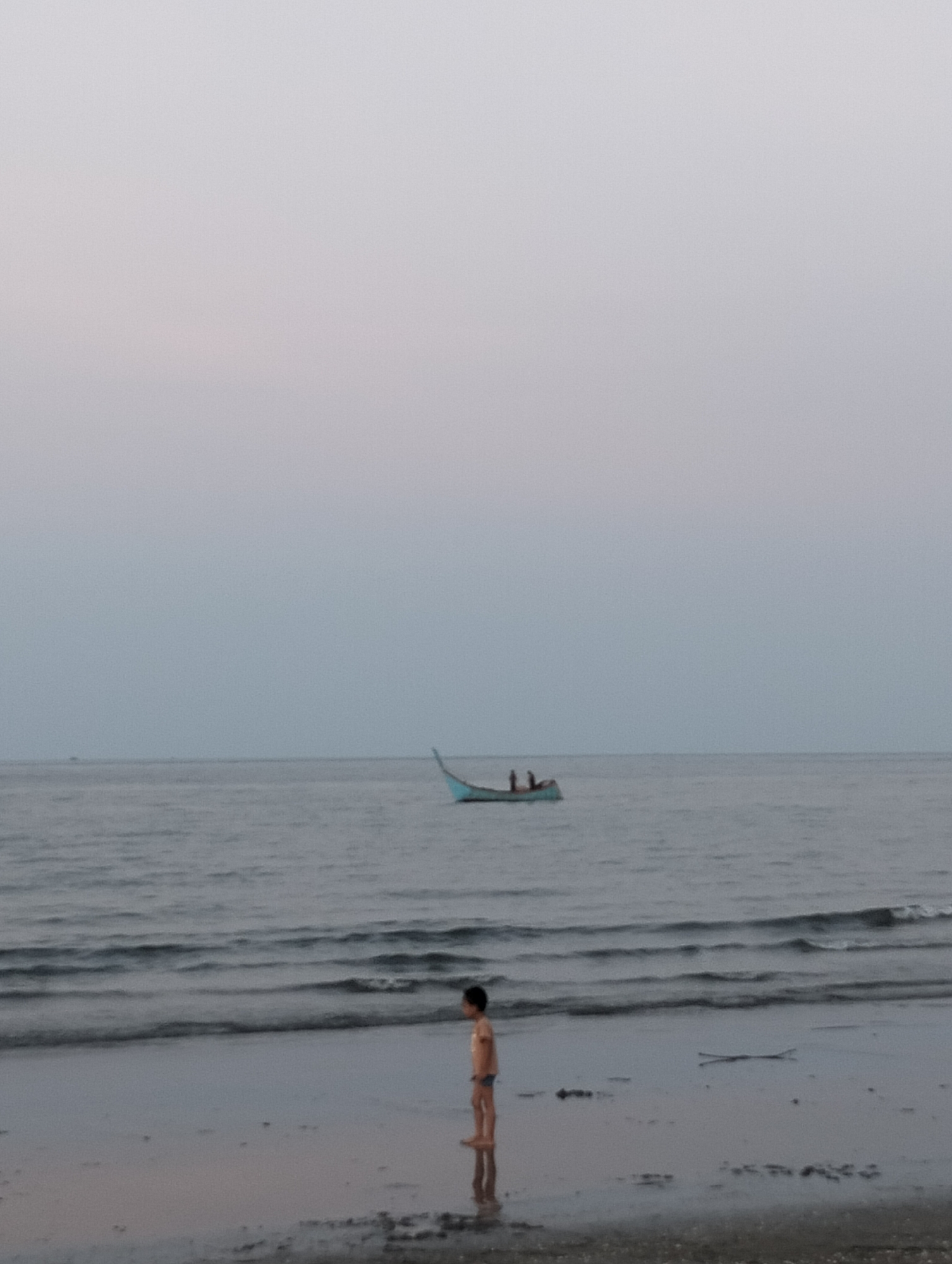 A photograph depicts a beach scene, likely at dusk or dawn, in Lhokseumawe, Indonesia. A young child stands on the wet sand near the water's edge, facing toward the ocean, with their reflection visible in the wet sand. The child is wearing minimal clothing. In the background, a small boat or canoe with two occupants is visible on the water. The sky is overcast, with a muted grey tone. The water appears calm, with gentle waves reaching the shore. A piece of driftwood lies on the sand near the child.