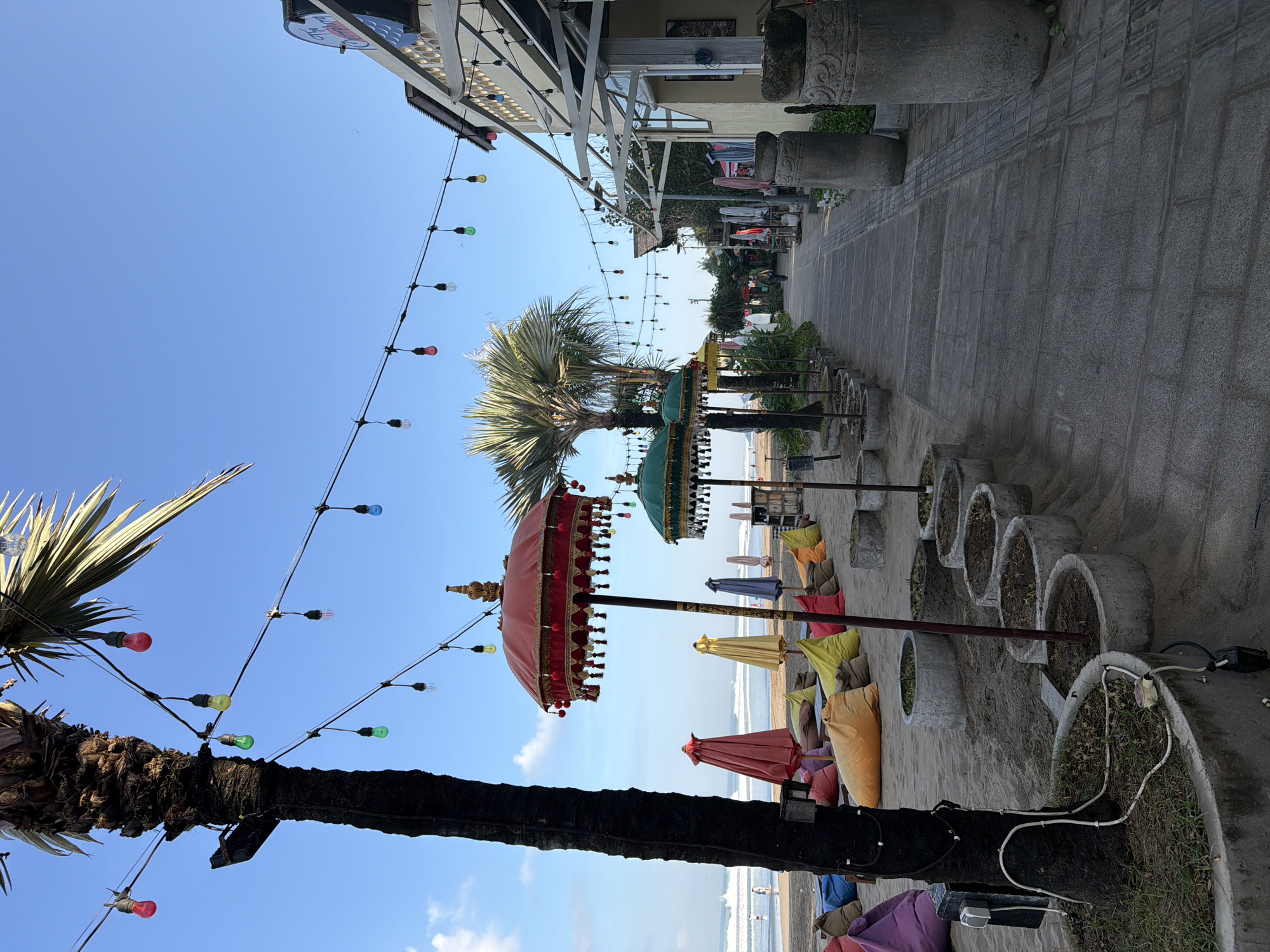 This image captures a vibrant, sunny beachside scene, likely in Kuta, Indonesia. The setting features a sandy area furnished with numerous colorful beanbag chairs in shades of red, yellow, orange, purple, and blue, suggesting a relaxed lounge or dining spot. Several traditional Balinese-style parasols (payung) with ornate fringes stand among the beanbags, colored red, green, yellow, and blue.
The scene is illuminated by strings of colorful Edison-style light bulbs (red, yellow, green, blue, and clear) strung across the sky, extending from palm trees and nearby structures. Two prominent palm trees, one with a dark trunk in the foreground, anchor the left side of the frame. Below these, a row of large, circular concrete planters, some containing sparse greenery, line the edge of the sandy area. Electrical wiring is visible on the ground connecting to these planters and structures.
To the right, a paved walkway made of square tiles runs parallel to the beach. Beyond the walkway, a series of buildings or businesses are visible, featuring awnings and some decorative stone elements or statues. One building has a partially visible sign that reads "The" followed by curved text, suggesting a bar or club name.
In the distance, the ocean stretches out under a clear blue sky with gentle waves breaking on the shore. A few very small, indistinct human figures can be seen on the beach near the waterline. The weather is bright and sunny, indicating daytime, possibly late afternoon, creating an inviting and leisurely atmosphere typical of a holiday destination. A small bird can be seen flying high in the clear sky.