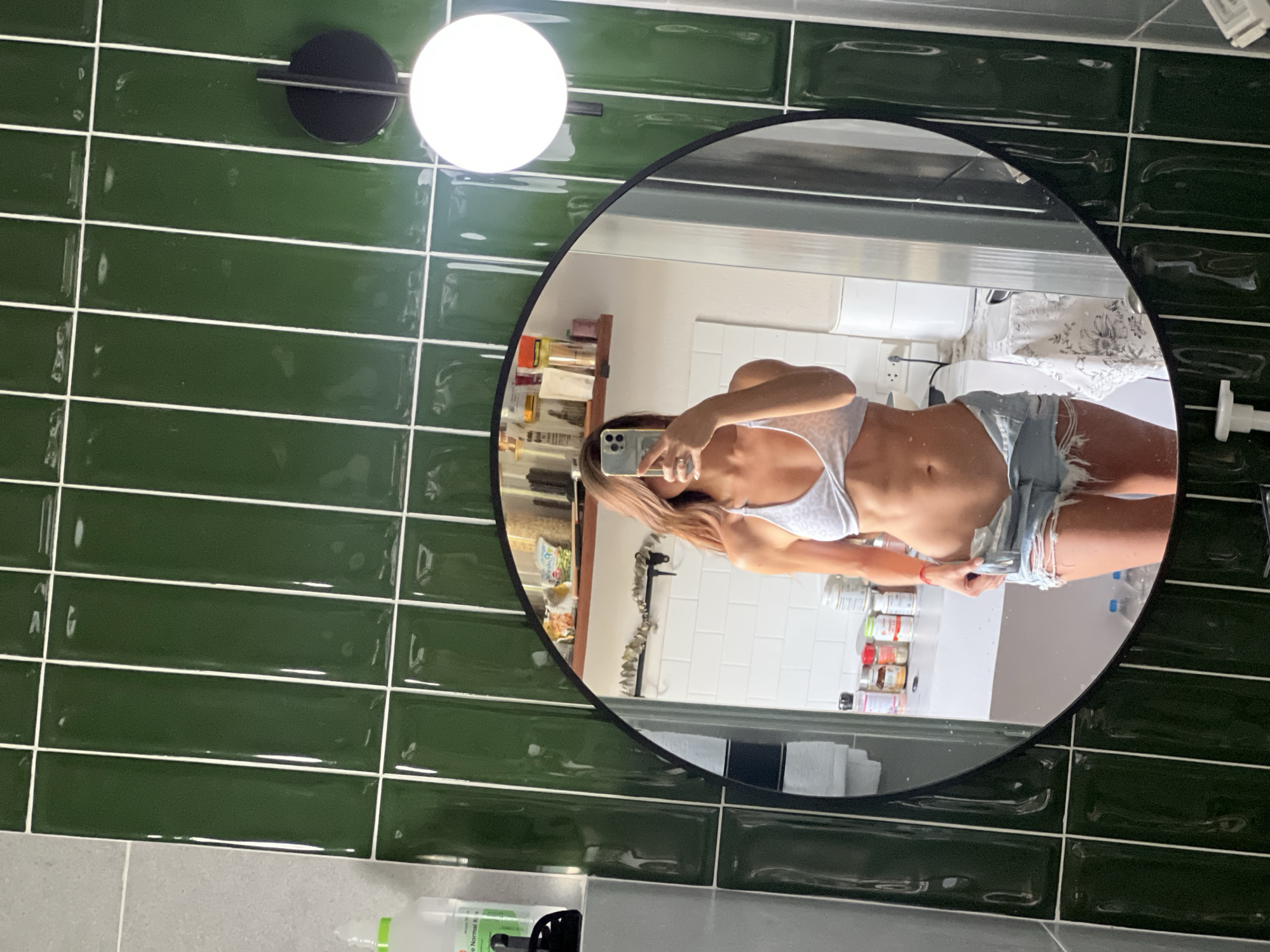 A female subject, visible from the chest to the mid-thigh, is captured in a mirror selfie within an unspecified indoor setting in the city of Unknown, Unknown. She has long, light brown hair partially obscuring her face and is holding a smartphone in her right hand. She wears a light-colored, subtly patterned halter-neck top and distressed light blue denim shorts, which are slightly pulled down by her left hand, revealing her toned abdomen. A red string bracelet is present on her left wrist. The selfie is taken in a large, round mirror with a black frame. The wall surrounding the mirror is clad in glossy, dark green rectangular tiles with white grout. To the left of the mirror, a grey tiled wall section is visible, along with a clear plastic bottle featuring a green cap and black pump. To the right, a wall-mounted black fixture with a spherical white light source is affixed to the green tiles. The reflection within the mirror shows a white tiled background, a white countertop holding numerous spice jars, supplement bottles, and cosmetic tubes, and a wooden shelf with additional containers. A white kitchen appliance and two large clear water bottles are also visible in the reflection.