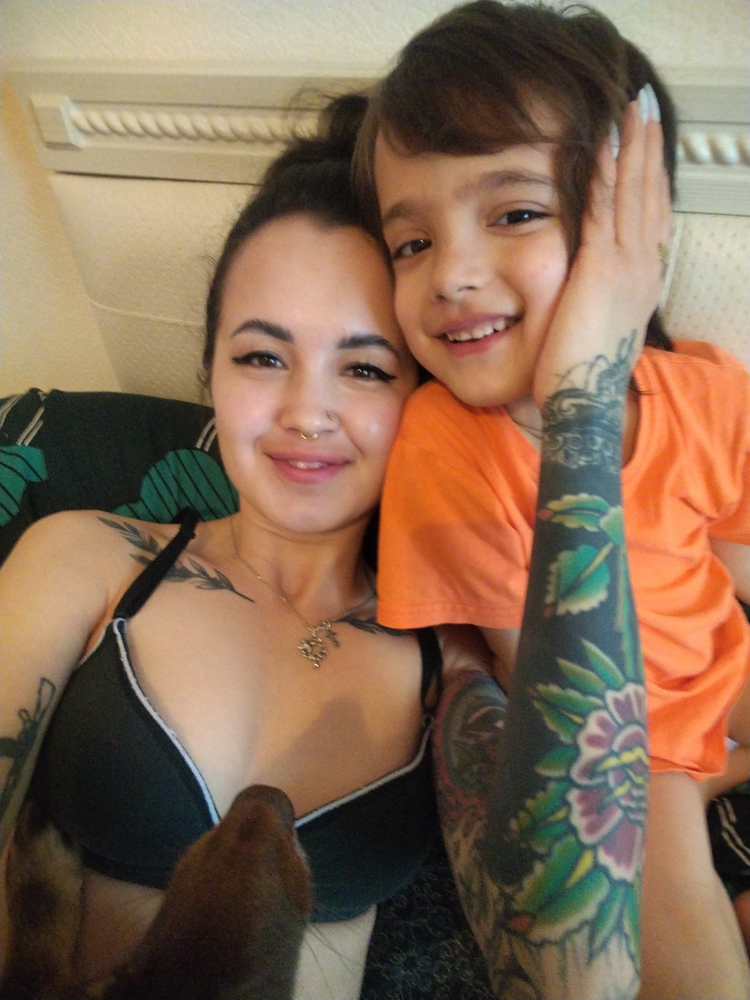 A woman and a child are posing for a selfie. The woman has dark hair in a messy bun, tattoos covering her right arm, a nose piercing, and is wearing a black bra with a white trim and a silver necklace. The child has brown hair and is wearing an orange t-shirt. The woman is touching the child's face with her hand. A brown dog is partially visible in the lower center of the frame. They are in a room with a white headboard. There are green patterned pillows behind them. The location is not in Noviy, Russia.