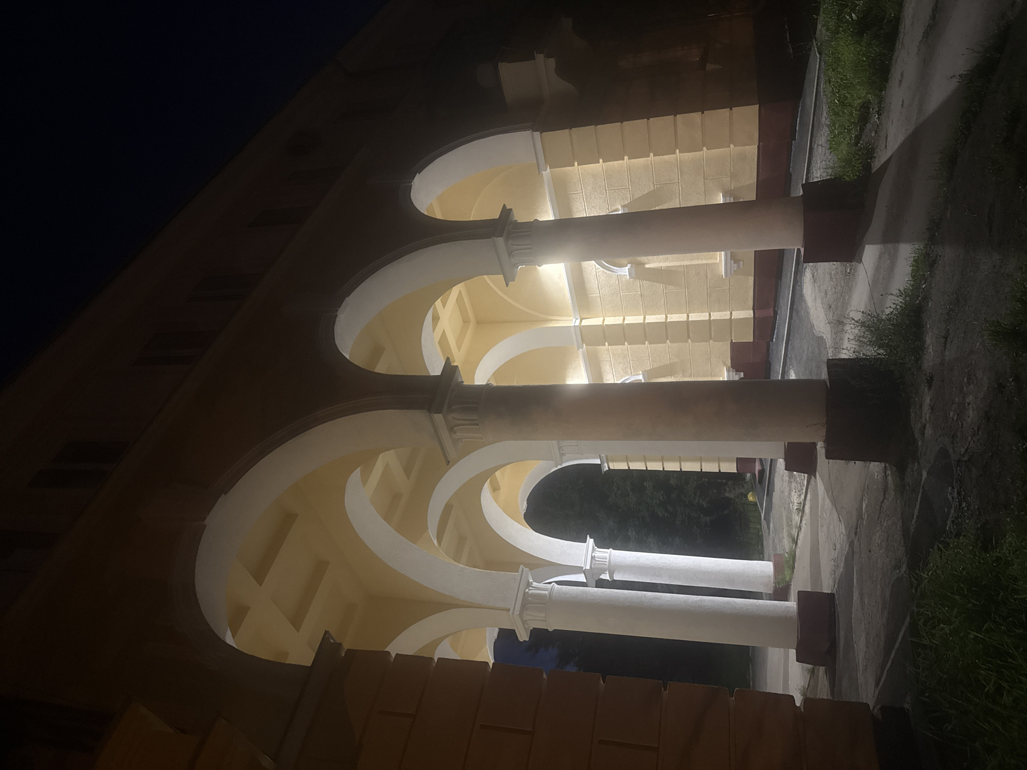 This nighttime photograph captures an architectural arcade, possibly part of a building in Magnitogorsk, Russia. The arcade features a series of arched openings supported by large, white cylindrical columns with decorative capitals. The facade behind the columns is painted a pale yellow with visible brickwork or textured plaster. The ceiling of the arcade is also yellow and features a grid-like pattern. The ground beneath the arcade is paved with a rough, uneven surface, and patches of grass are visible on the left and right sides of the frame. The scene is dimly lit by artificial lights, casting long shadows from the columns onto the ground. The sky above is dark, indicating it is night. No people or vehicles are present in the image.