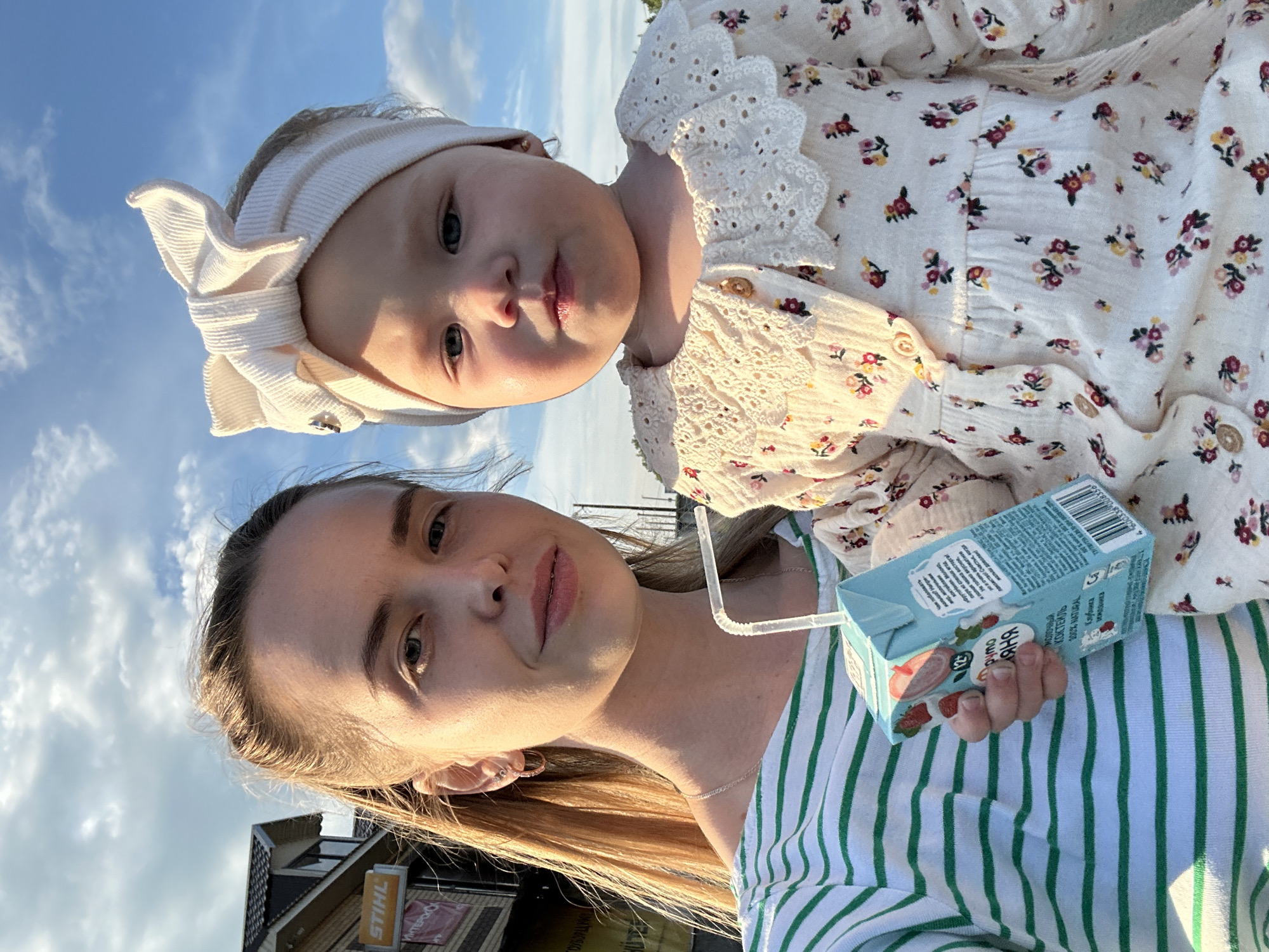 A woman and a young child are standing outside with a building and cloudy sky in the background. The woman is on the left side of the frame, wearing a green and white striped shirt and holding a small juice box with a straw. She has blonde hair pulled back and is looking at the camera. The child is on the right side of the frame, wearing a white floral dress with a large white bow headband. The child is looking at the camera with a neutral expression. The building in the background appears to be a commercial establishment with signage. The lighting suggests it is daytime, likely late afternoon or early evening due to the warm glow. The overall impression is a casual, candid moment captured outdoors. The location is identified as Azinsky, Russia.
