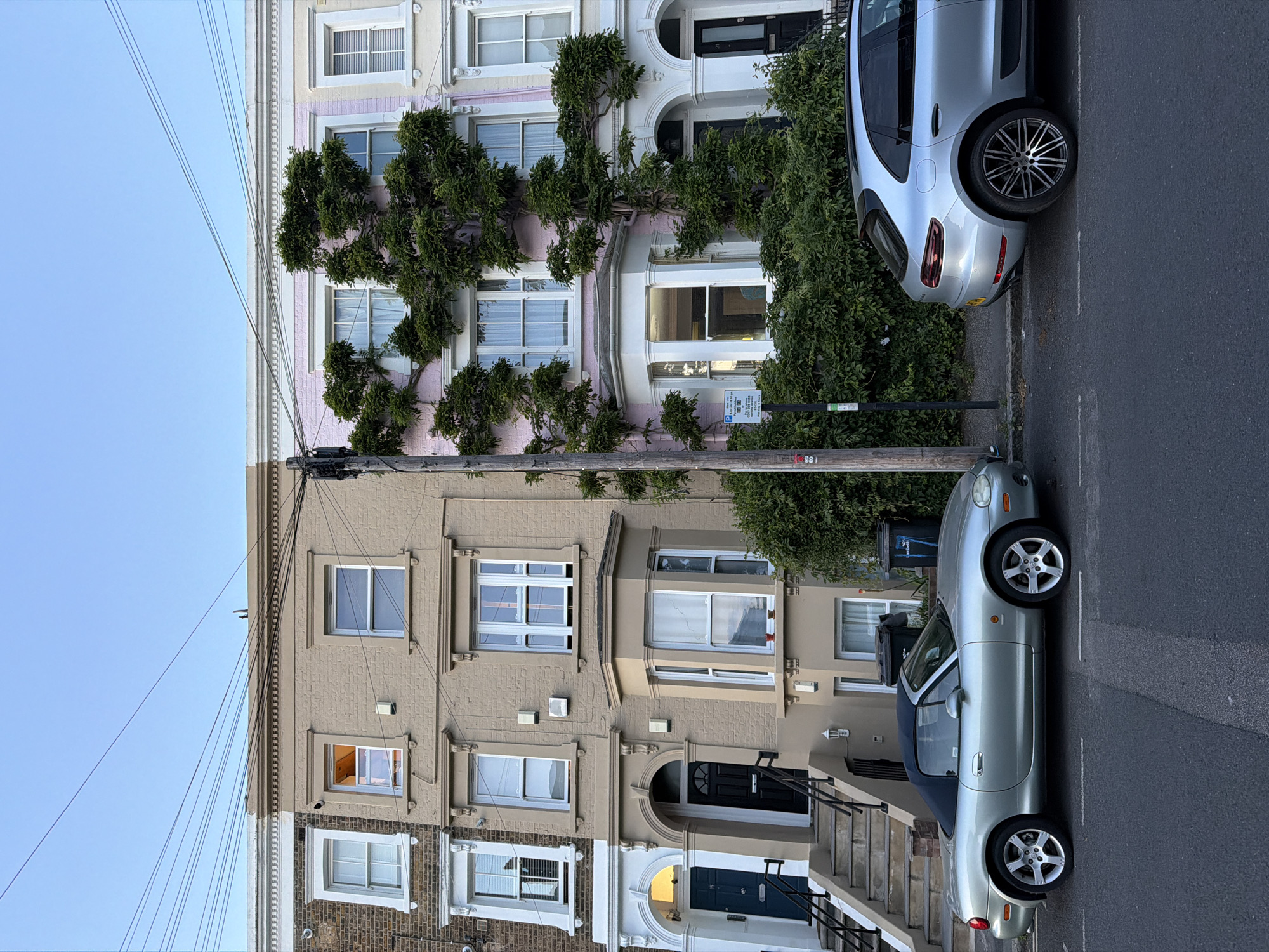 The image depicts a street scene in London, United Kingdom, showing a row of terraced houses with a light blue sky visible above. The houses exhibit variations in facade colors and architectural details, including different window styles, doors, and exterior paint. One house is painted pink, another is beige, and the lower one is partially brick and beige. A vine or climbing plant is growing on the facade of the pink house. Two silver-colored cars are parked along the curb. There are visible utility poles and power lines in the upper left of the frame. The street itself appears to be asphalt. The overall impression is a typical residential street in a London neighborhood.