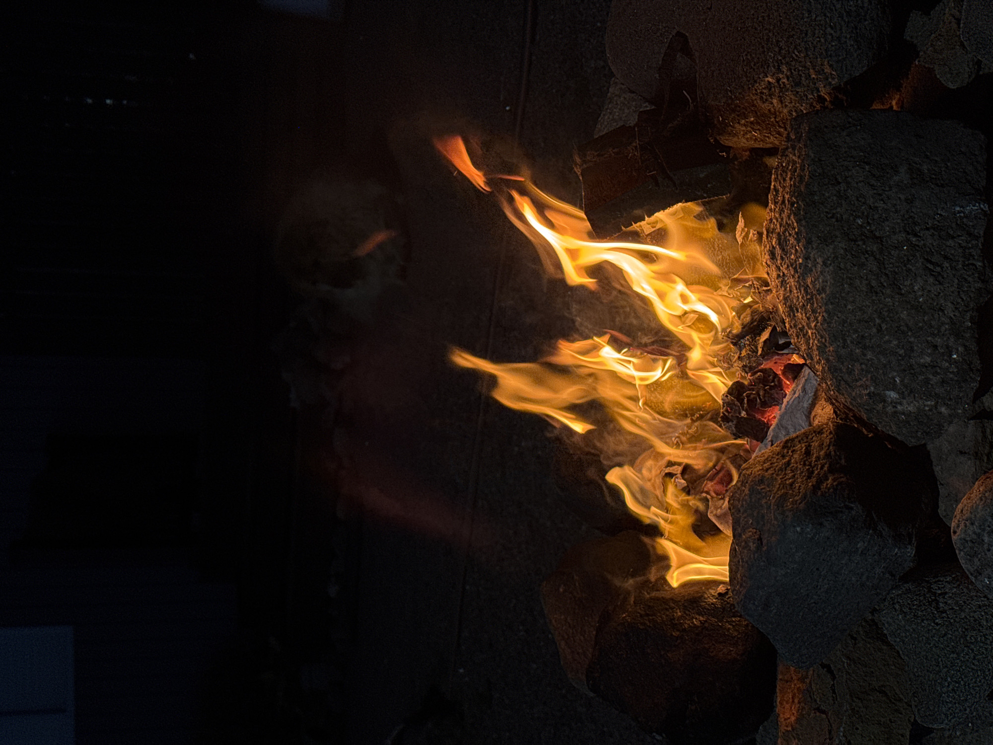 A campfire burns in a rocky pit at night. The flames are bright orange and yellow, casting a warm glow on the surrounding dark gray rocks. The background is black, suggesting it is a dark night. There are no people visible in the image. The scene appears to be outdoors, possibly in a park or campground. There is no indication of the specific location within Kempelevo, Russia.