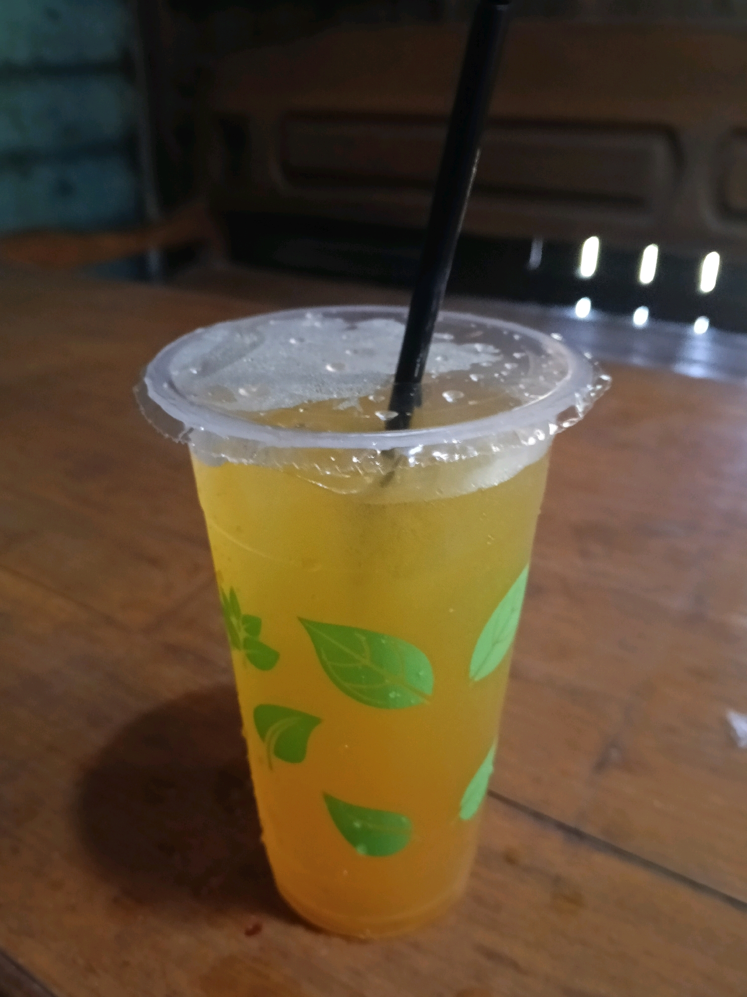 A close-up shot captures a translucent plastic cup filled with a yellow, iced beverage and a black straw. The cup is decorated with green leaf patterns and sits on a wooden surface, possibly a table. Condensation is visible on the outside of the cup, indicating it's cold. The background is blurry, showing what appears to be a wooden interior and some bright vertical lights. This image was taken indoors, likely in a cafe or restaurant, and there is no information to suggest it is specifically located in Nglungger, Indonesia.