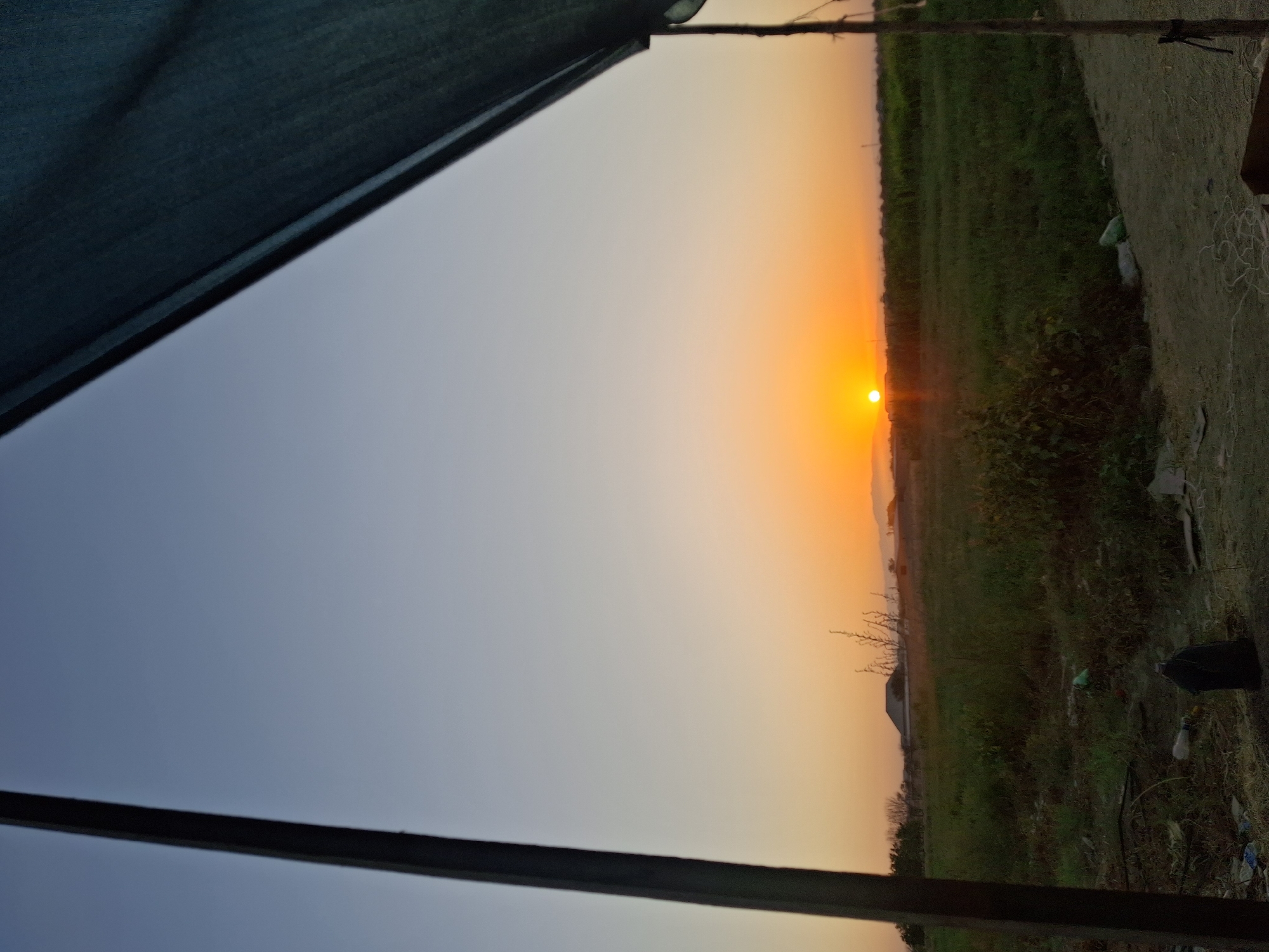 The image depicts a sunset scene with a clear sky transitioning from light blue to orange. In the foreground, a wooden pole and a dark fabric are visible on the right side, framing the view. A grassy field stretches out towards the horizon, with scattered vegetation and some debris on the ground. In the distance, a building with a pyramid-shaped roof can be seen, along with trees and the silhouette of mountains. The overall impression is one of a rural or semi-urban landscape at dusk in Utmayli, Uzbekistan.