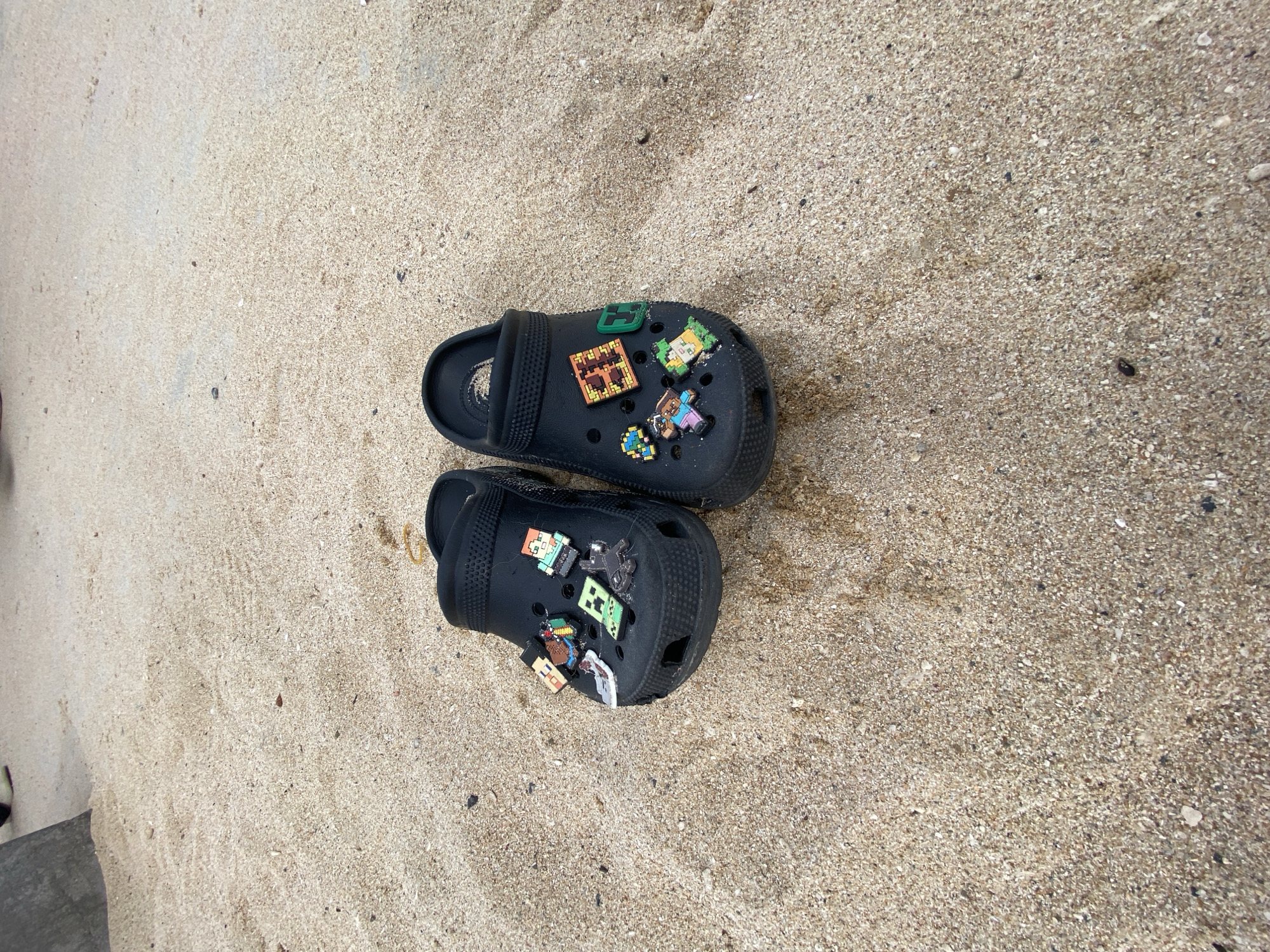 A pair of black Crocs adorned with pixelated charms, resembling characters and objects from the video game Minecraft, rests on the sandy shore of a beach. The sand is light brown and speckled with small shells and dark debris. The Crocs are positioned side-by-side, with the left shoe closer to the camera. In the background, a dark, textured object, possibly a rock or a piece of driftwood, is partially visible on the left side of the frame. The overall scene suggests a casual setting, likely a recreational area on the coast. This is a generic beach scene, and it is not possible to determine the specific location within Nha Trang, Vietnam, based solely on the visual information provided.